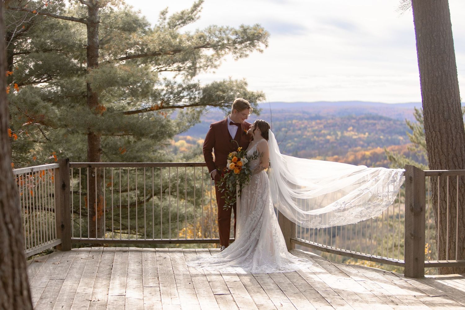 A couple embraces on a scenic mountain overlook deck with autumn foliage and rolling hills in the background.