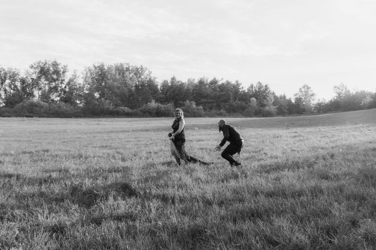 Two silhouetted figures play and run together across a grassy hilltop against an overcast sky.