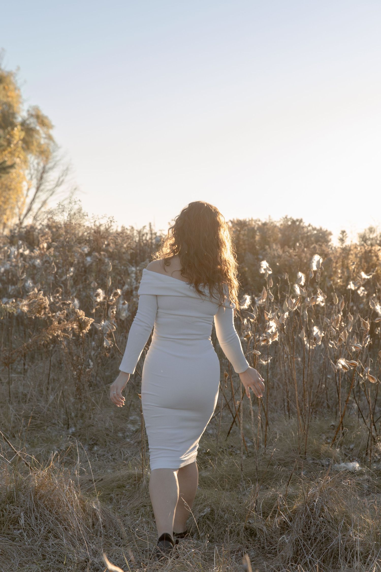 A person in a white fitted dress walks through a field of tall grass and wildflowers at sunset.
