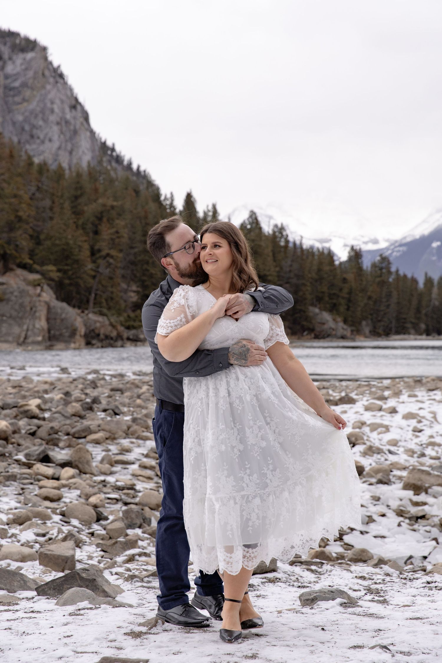 A romantic moment captured by a mountain river with snow and evergreens creating a scenic winter backdrop.