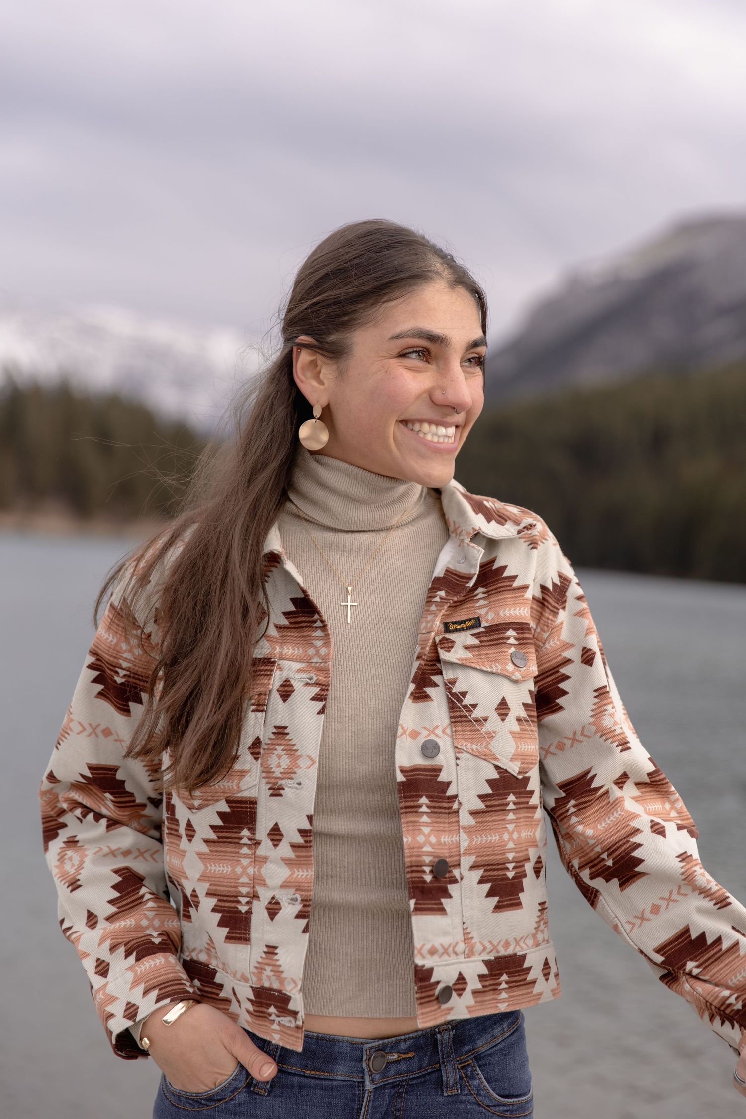 Woman in patterned jacket and turtleneck poses playfully by mountain lake on cloudy day.