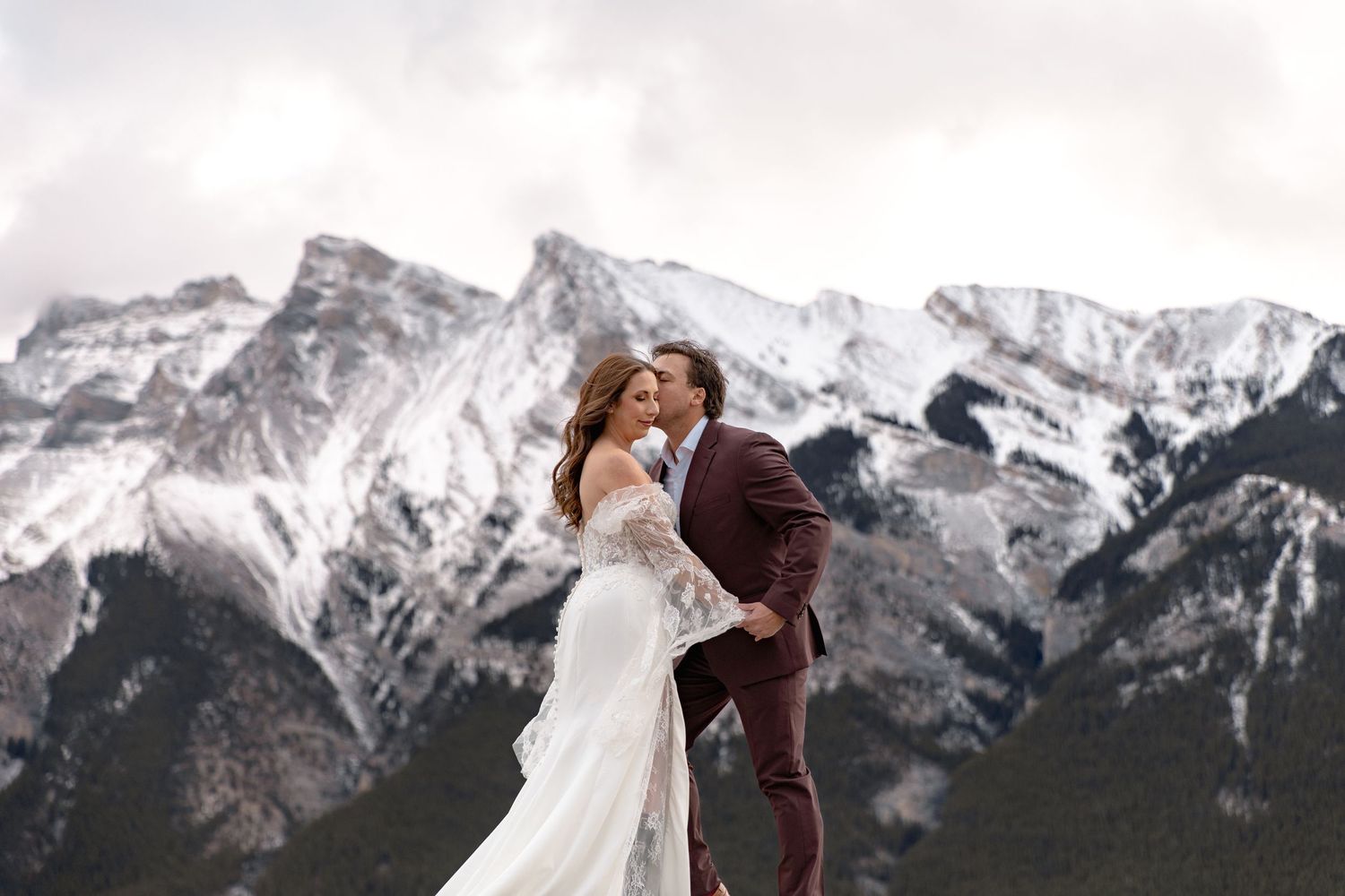 A couple in wedding attire embrace while standing against a backdrop of snow-covered mountain peaks in winter.