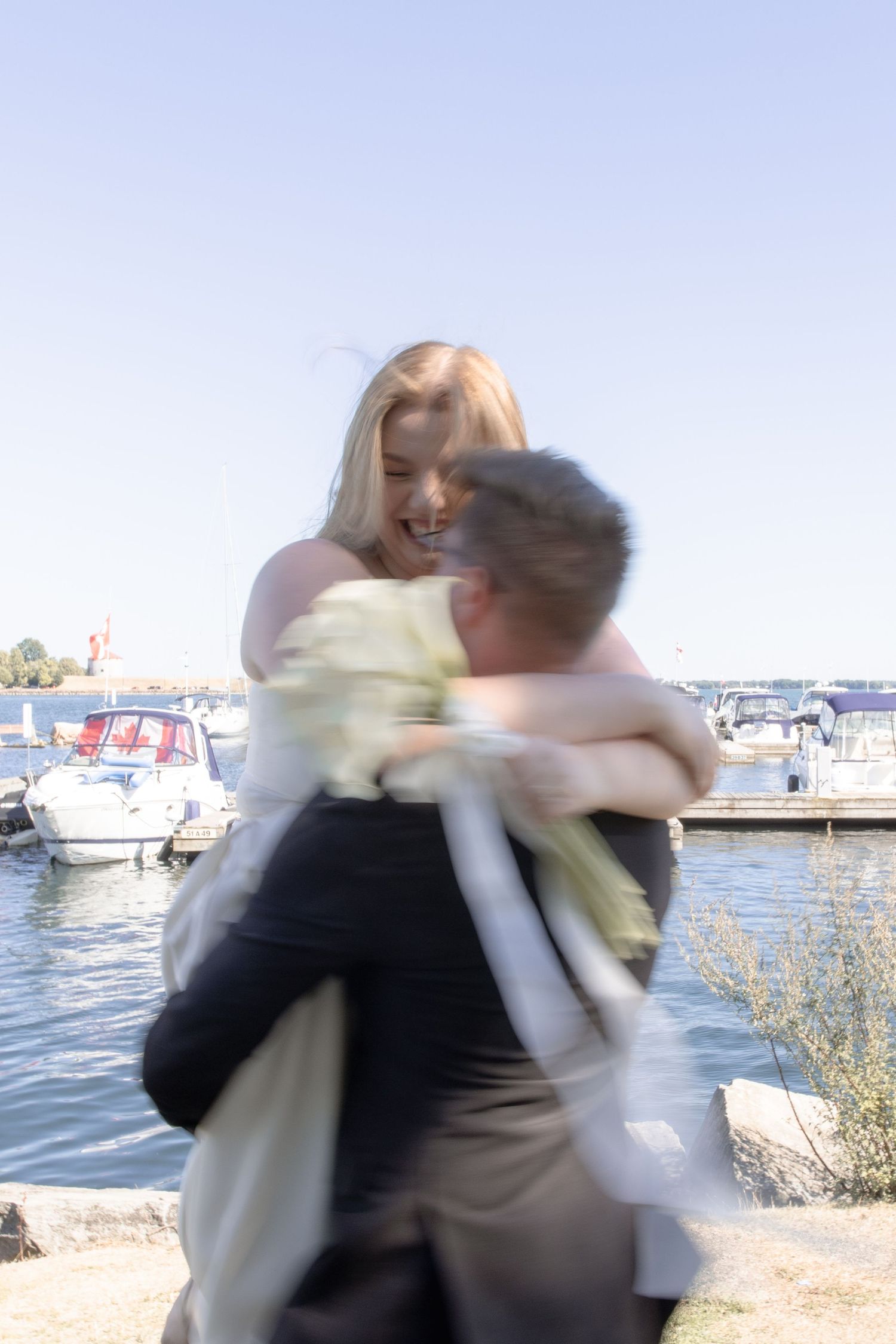 Two people in formal attire embrace and spin joyfully by a marina with boats in the background.