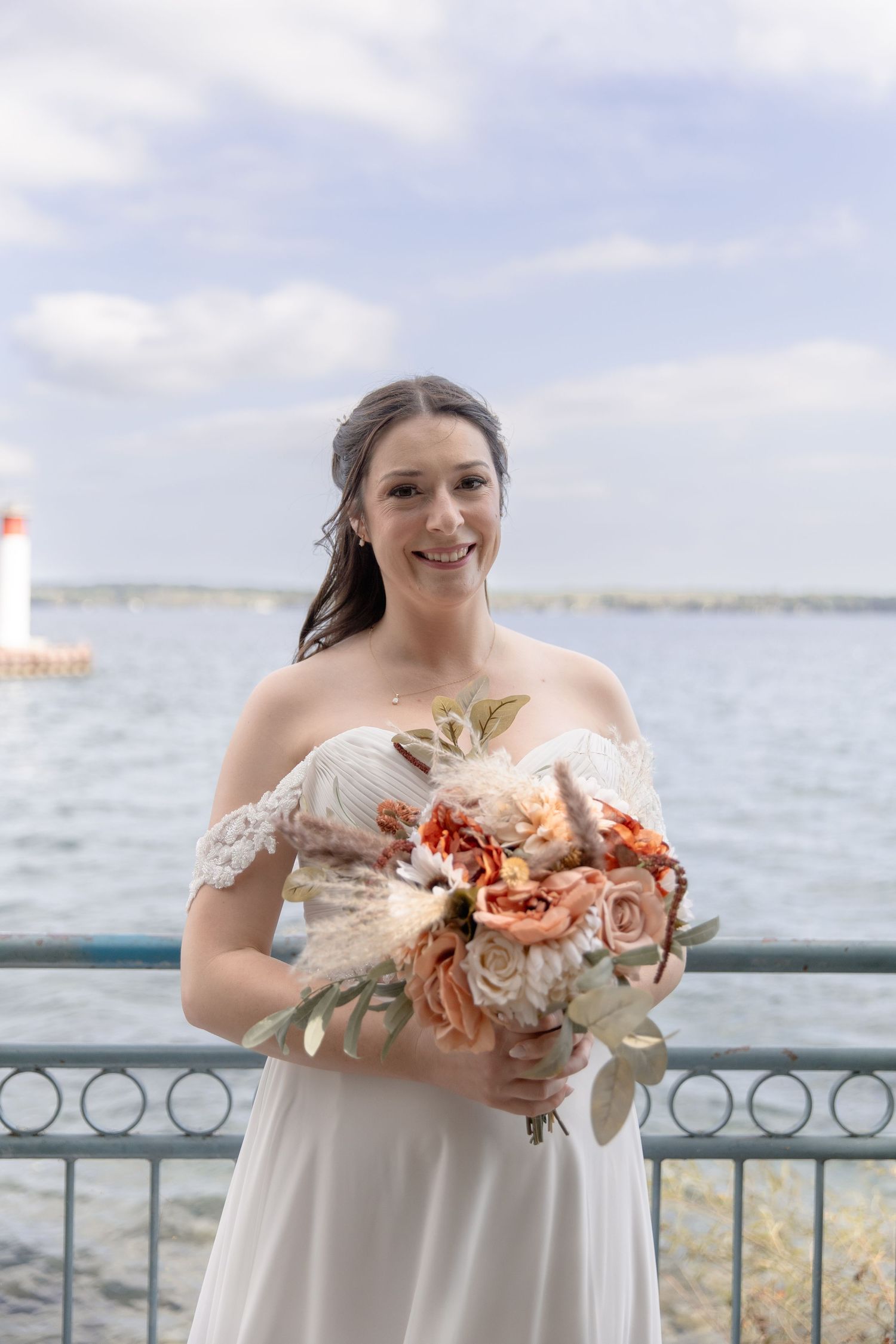 Bride in off-shoulder white dress holds peach and coral floral bouquet against waterfront backdrop.