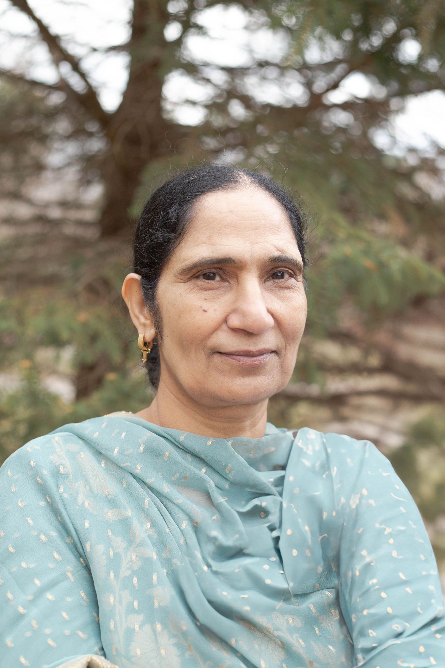 A person wearing a light blue patterned dupatta smiles gently while posing outdoors with trees in the background.