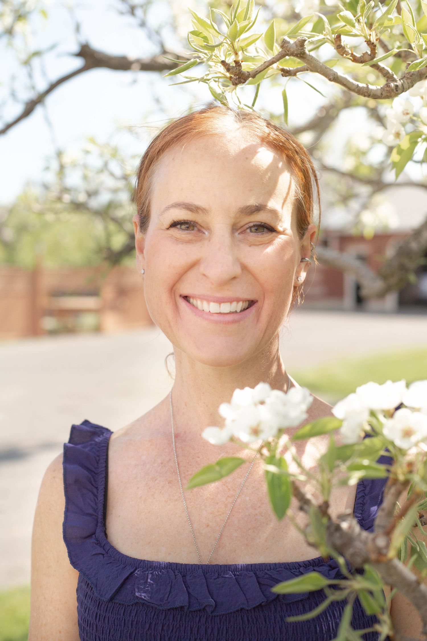A person in a navy blue ruffled top poses outdoors on a sunny day with white flowers and olive branches in the background.