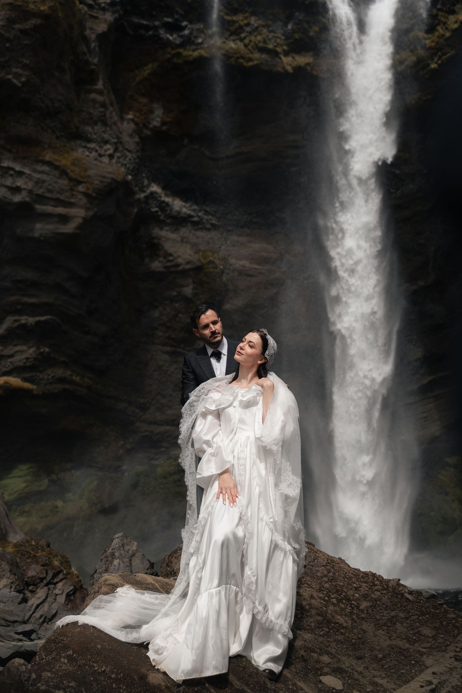 Bride in flowing white gown and groom in black tux pose romantically before a majestic waterfall on rocky terrain.