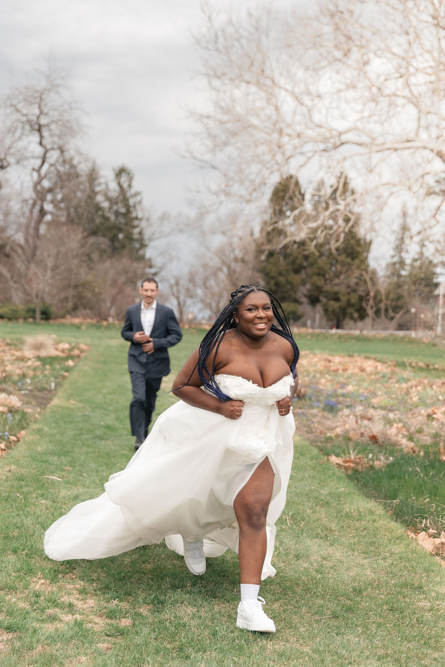 Joyful bride in white gown runs toward camera while groom follows behind in lush garden setting.