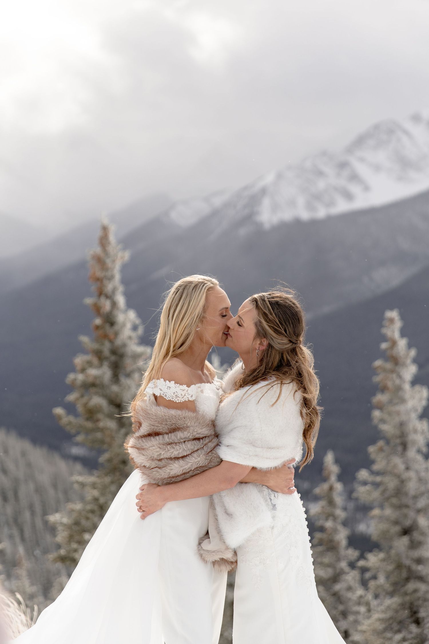A romantic winter mountain scene shows two people in white sharing an intimate moment among snow-covered pines.