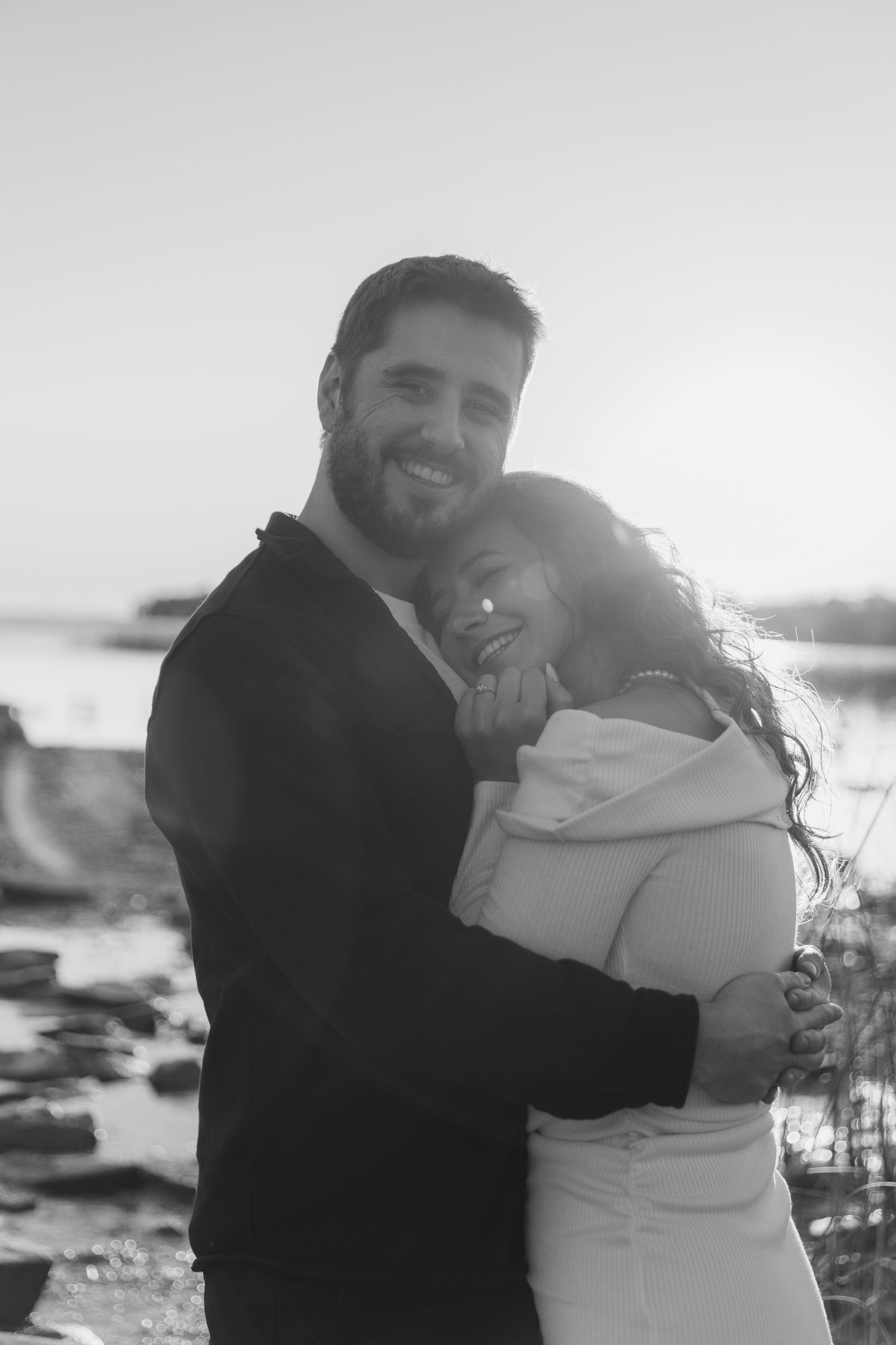 A romantic black and white portrait of a couple embracing on a rocky beach at sunset.