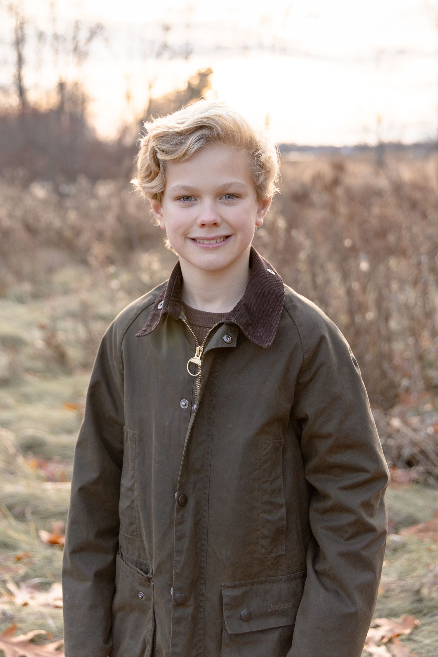 Young person in olive green jacket poses outdoors on an autumn day with golden light and fallen leaves in background.