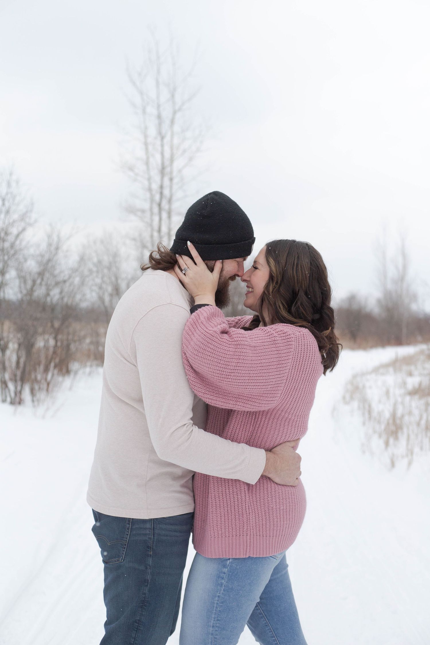 Intimate winter engagement photo of couple kissing on snowy path.