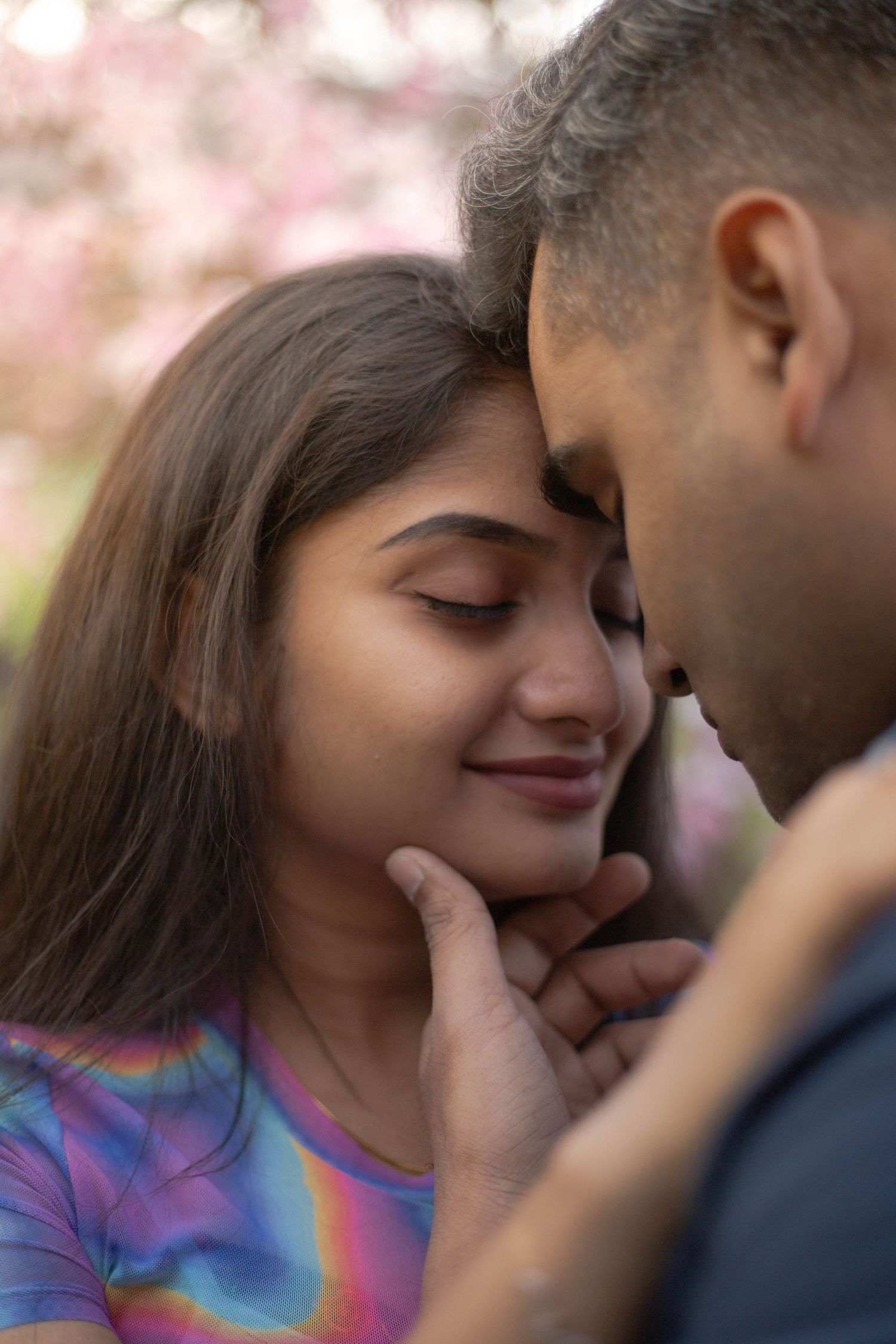 Intimate couple shares tender moment among cherry blossoms while wearing colorful tie-dye clothing.