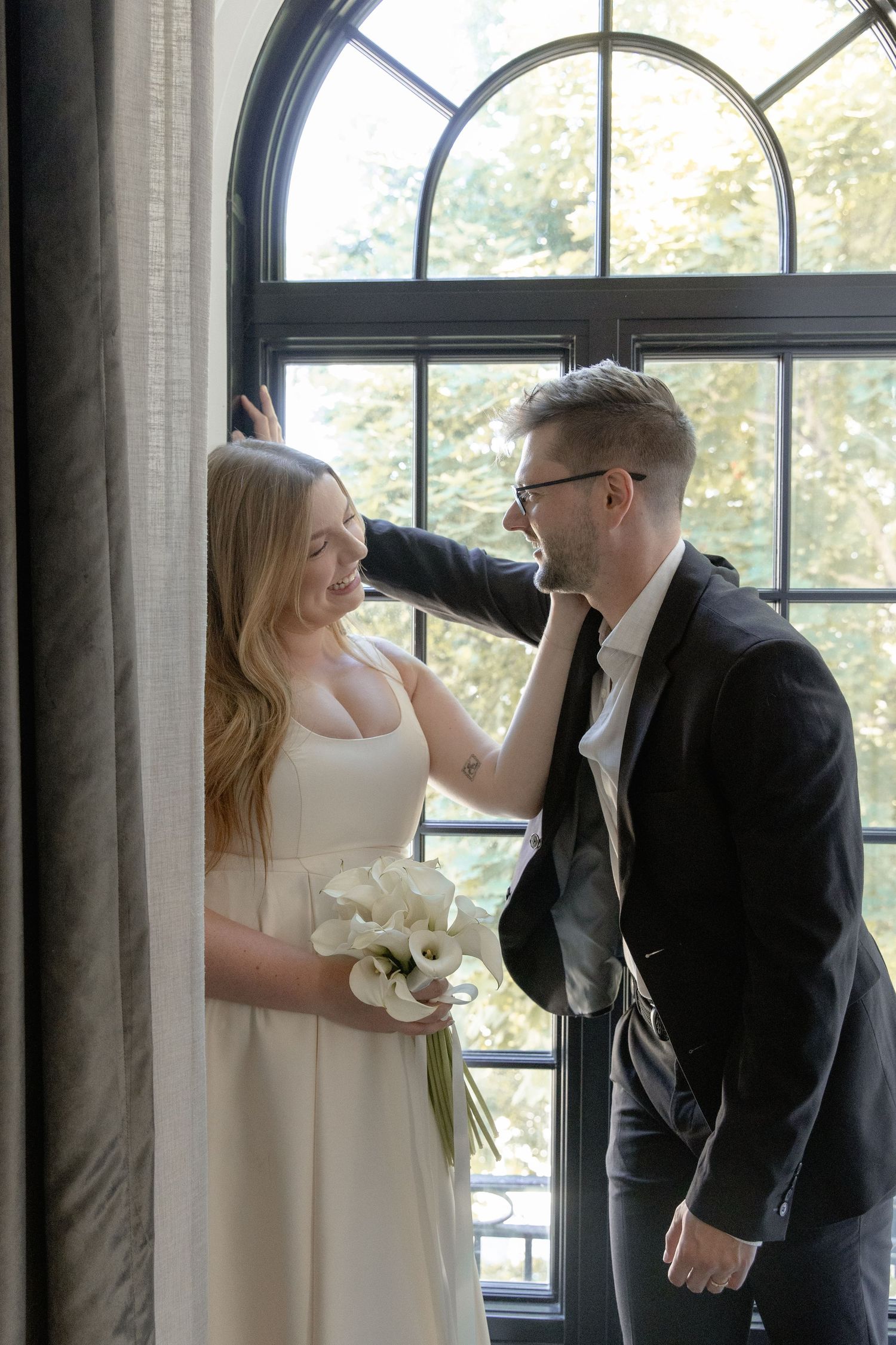 A romantic wedding couple shares a tender moment by a large arched window while the bride holds a white bouquet.