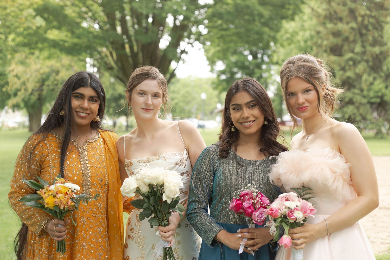 Four friends stand together outdoors holding colorful bouquets of flowers at a special event.