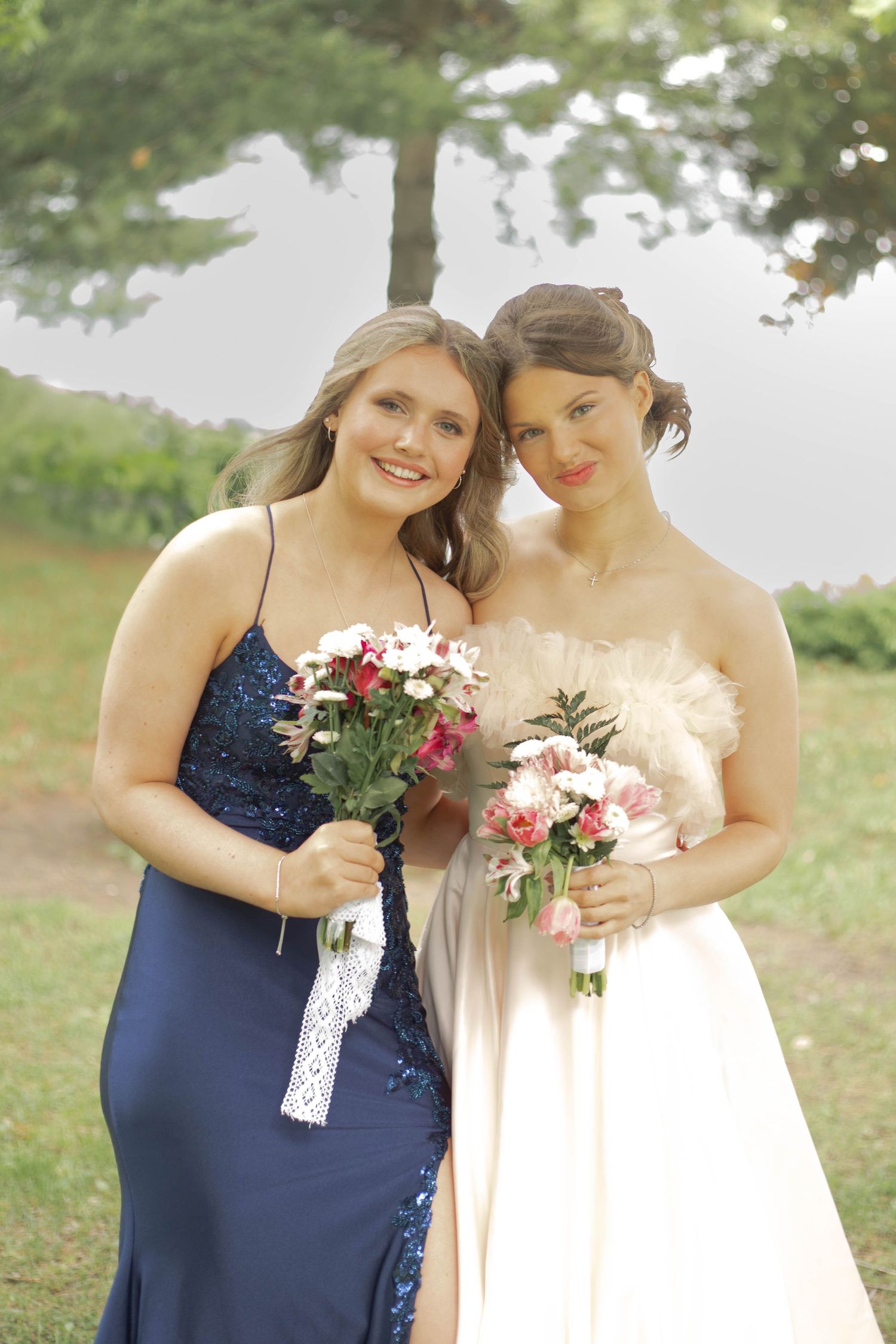 Two people in formal dresses stand together outdoors holding floral bouquets against a nature backdrop.
