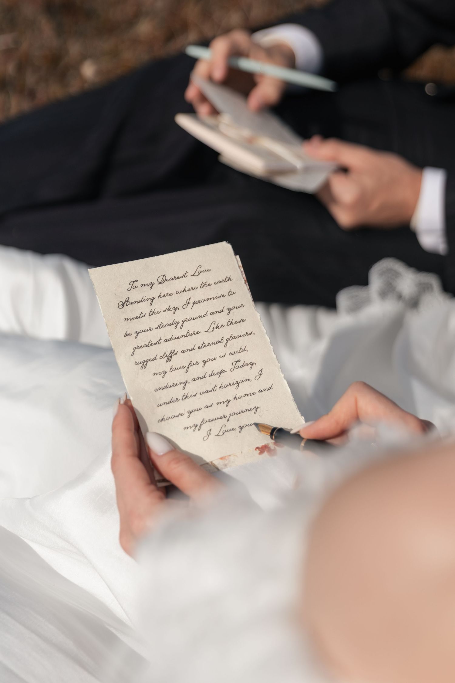 Bride and groom reading handwritten wedding vows on aged paper during outdoor ceremony.