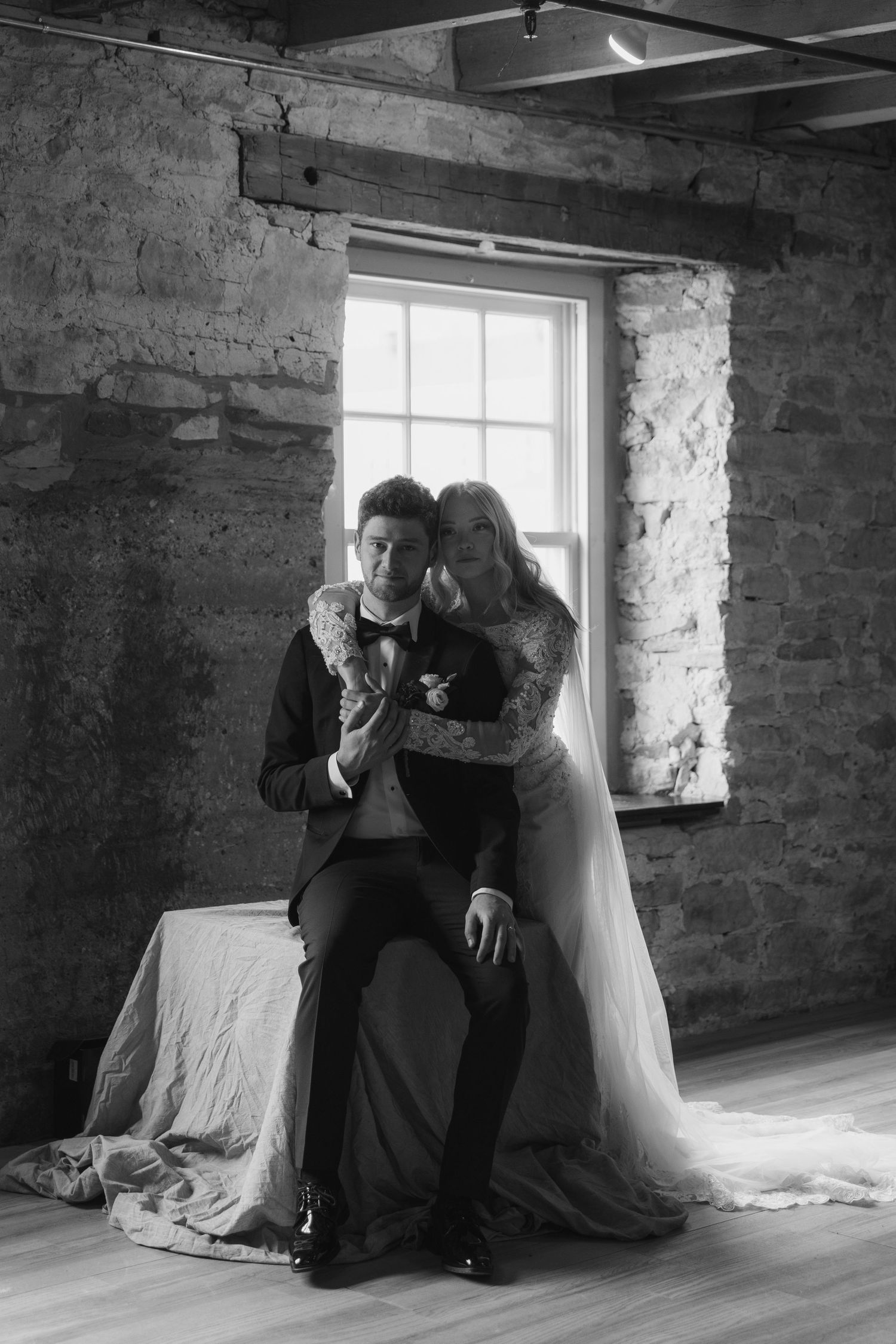 A couple in wedding attire share an intimate moment by a rustic stone wall window.