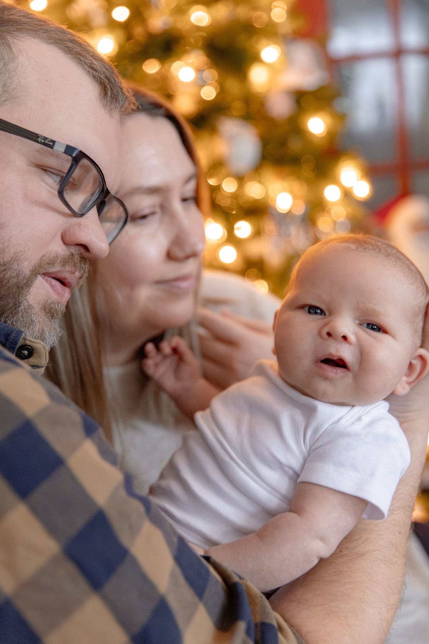 Heartwarming holiday scene with baby and parents sharing joyful moments against twinkling Christmas tree lights in background.