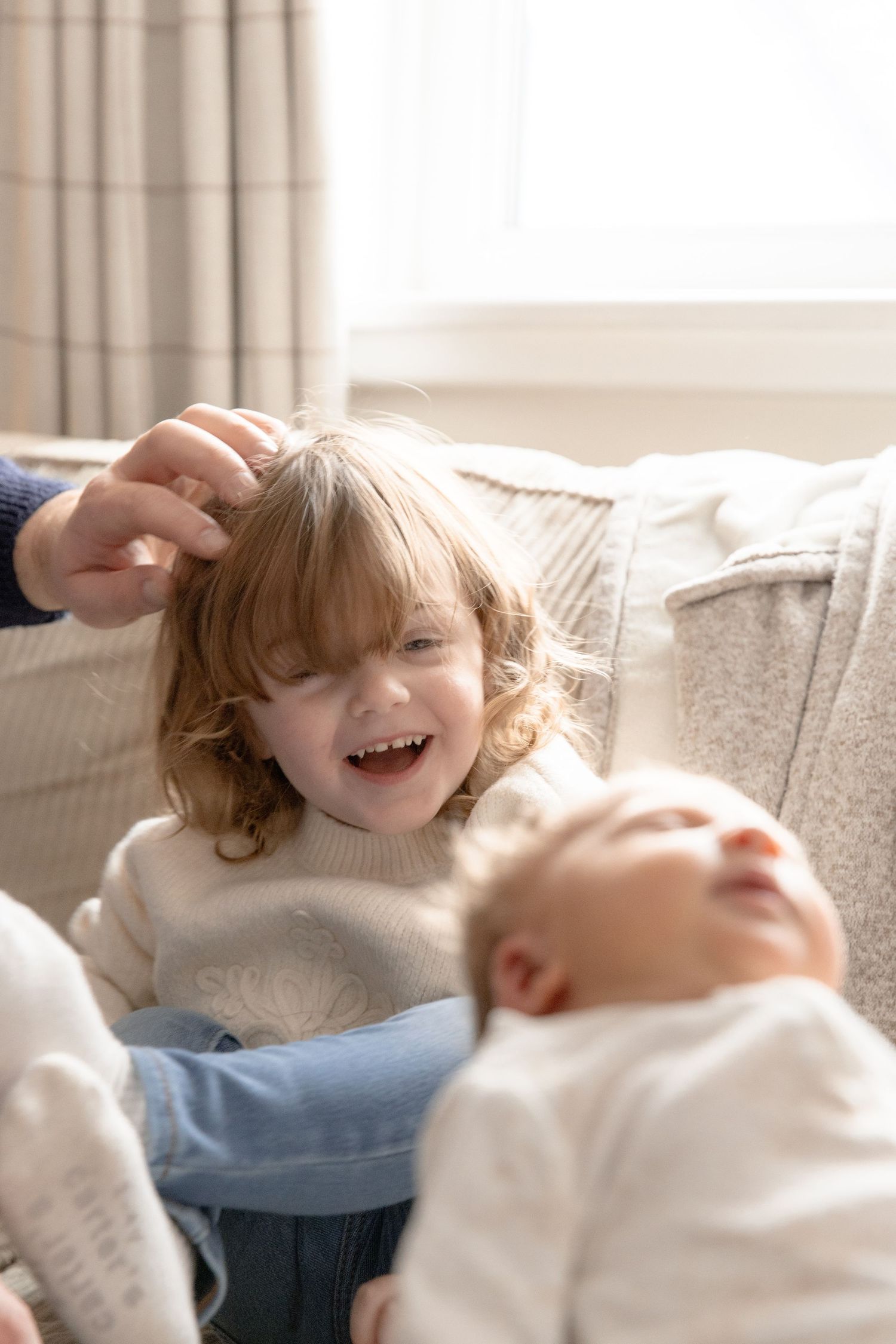 Two children share a joyful moment while relaxing together on a couch in a sunlit room.