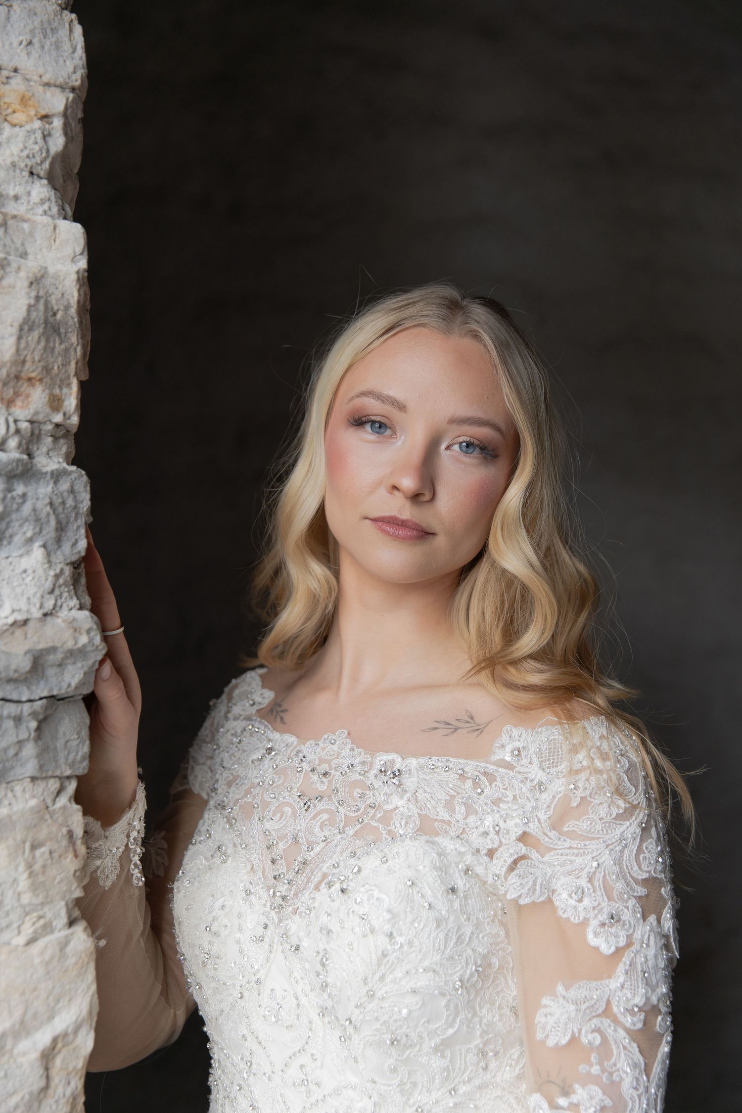 Elegant white lace wedding dress with intricate details photographed against a dark stone wall.