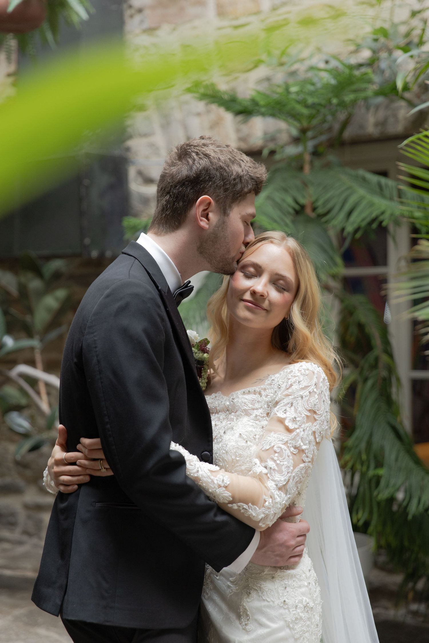 Wedding couple embracing in an intimate moment surrounded by lush greenery and stone architecture.