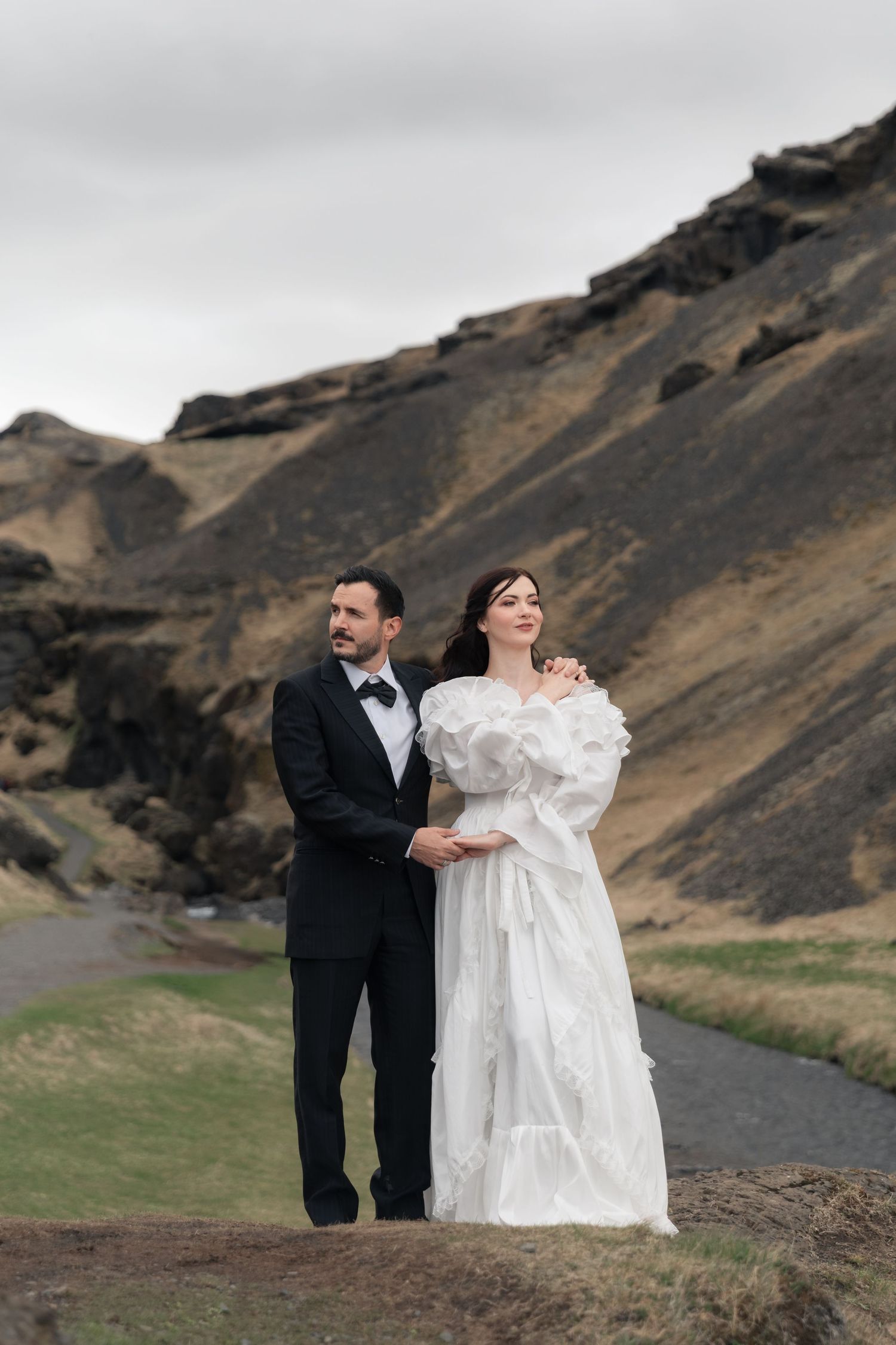 Bride and groom pose in Iceland's dramatic volcanic landscape, she in ruffled white gown, he in classic black tuxedo.