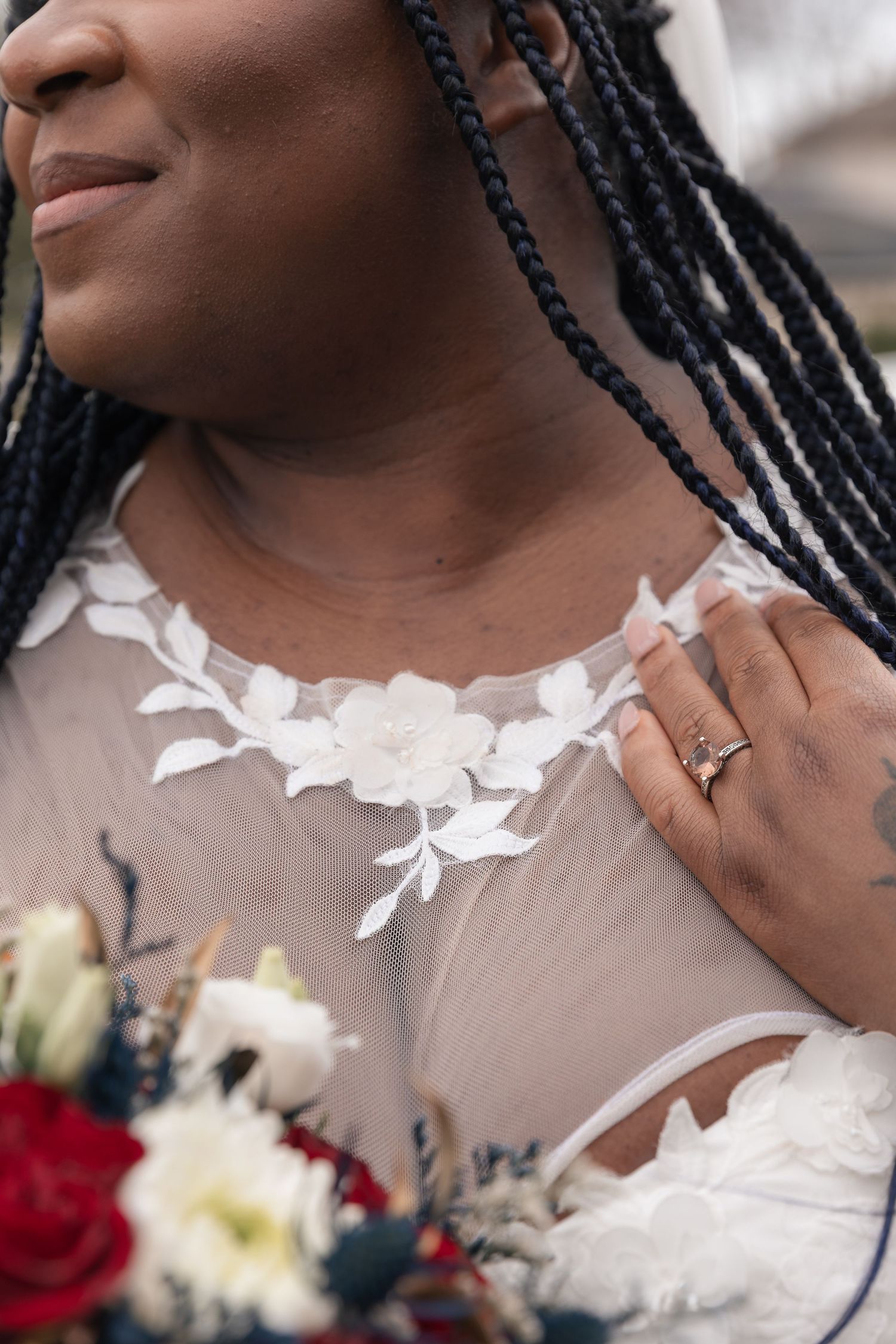 Close-up of a Black bride in a sheer floral lace wedding dress, holding a red and white bouquet, wearing a gold ring.