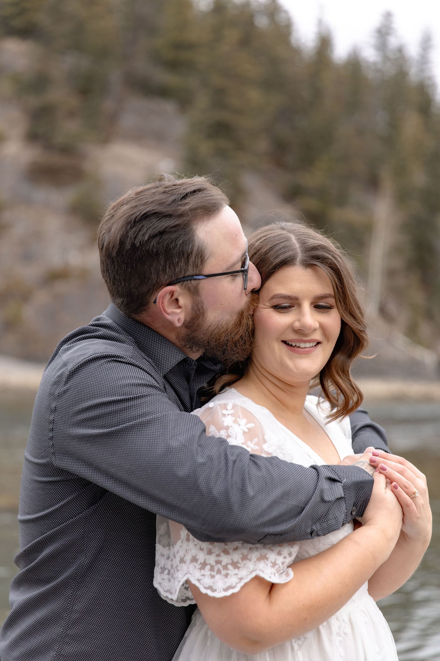 A couple shares an intimate moment by a lake with evergreen trees in the background during their wedding photoshoot.