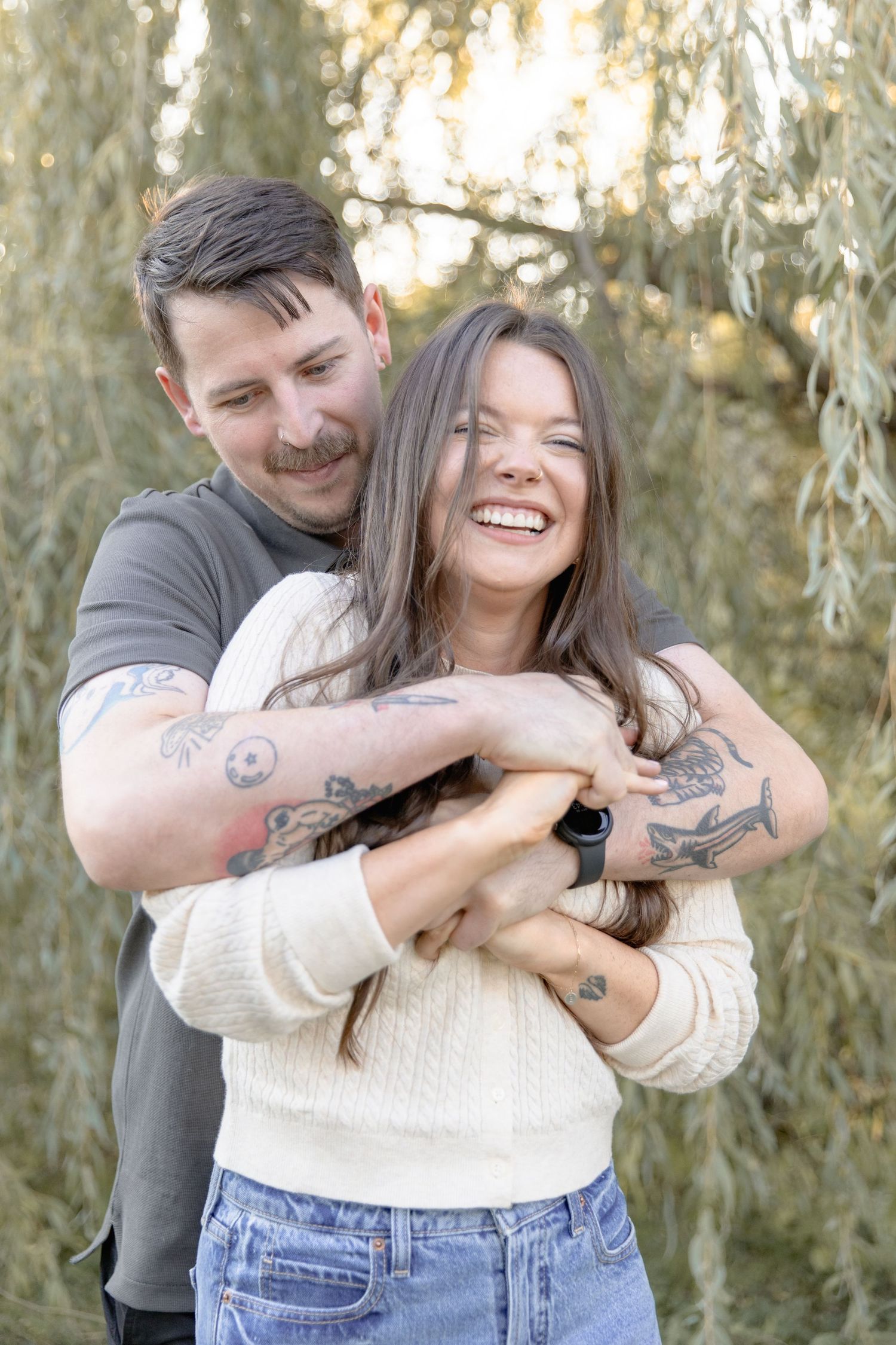 A playful couple laughs and embraces while posing against a natural outdoor backdrop.