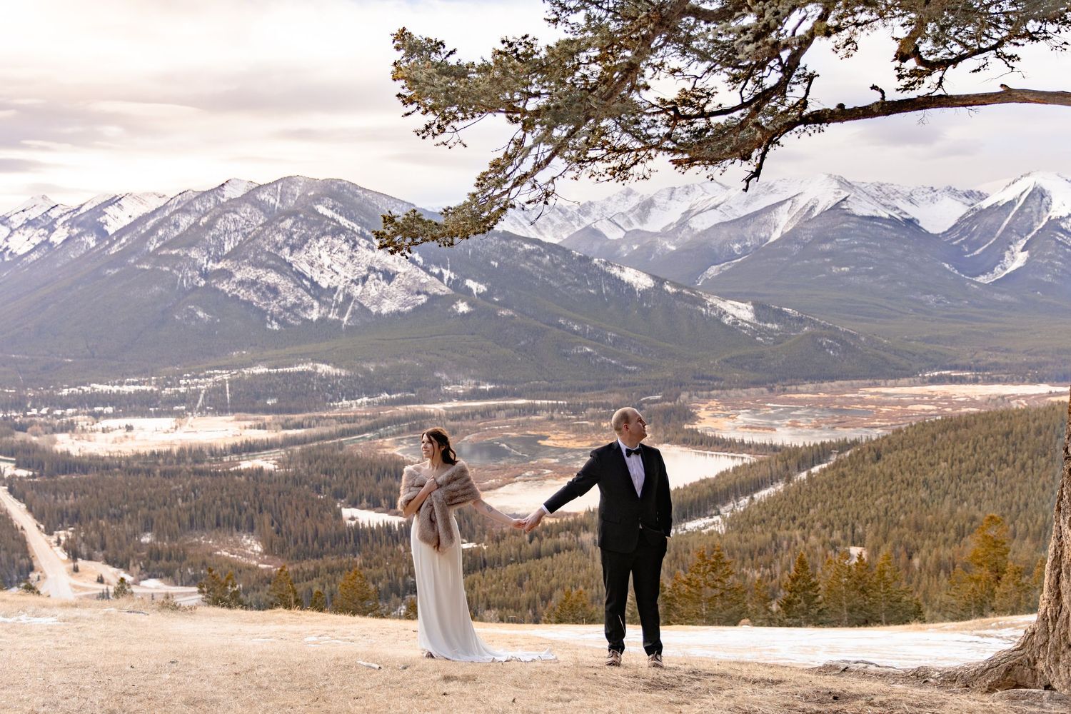 A winter wedding ceremony takes place on a mountain overlook with snow-capped peaks and evergreen forests in the background.