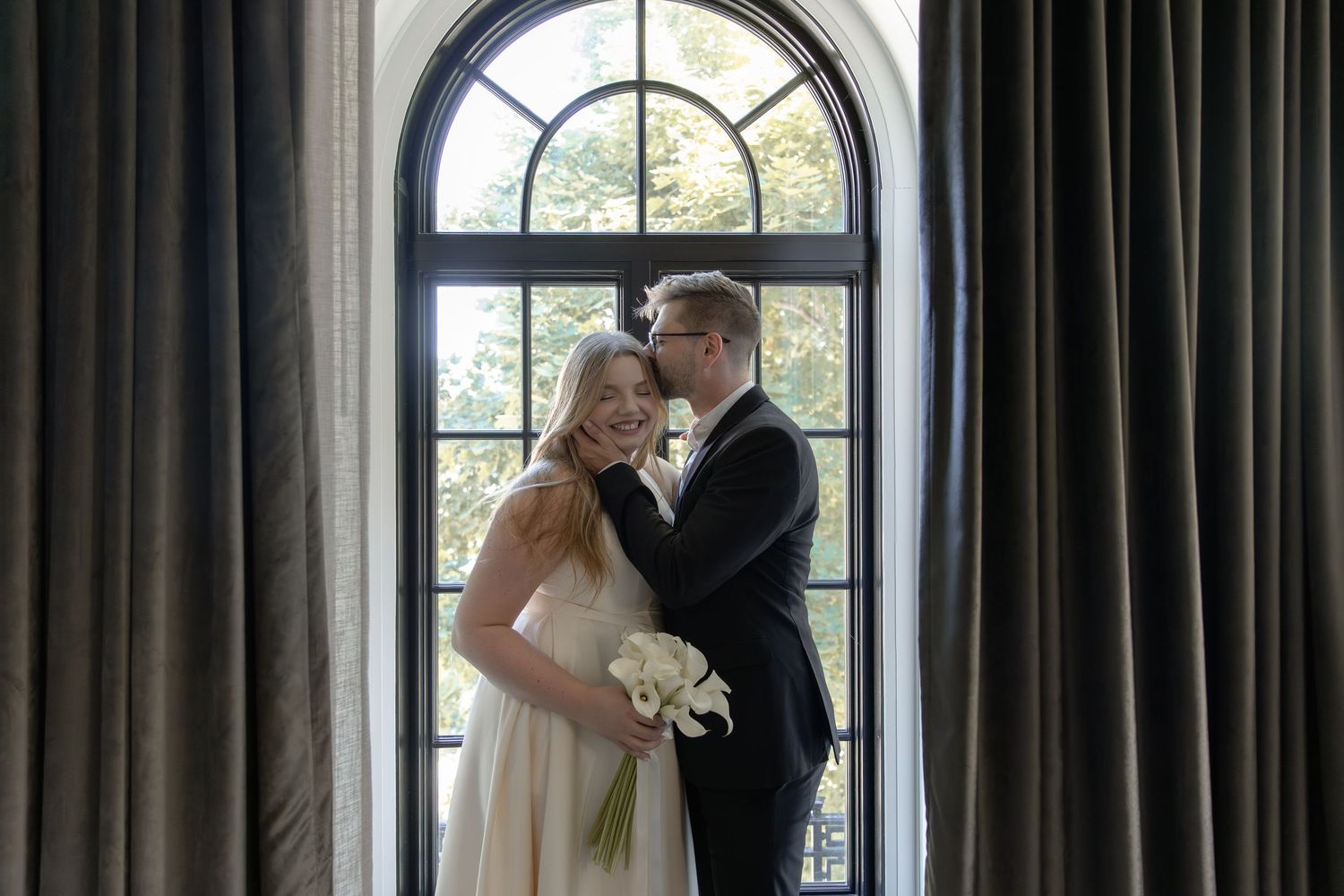 Newlywed couple embraces romantically beneath grand arched windows in a luxurious venue.