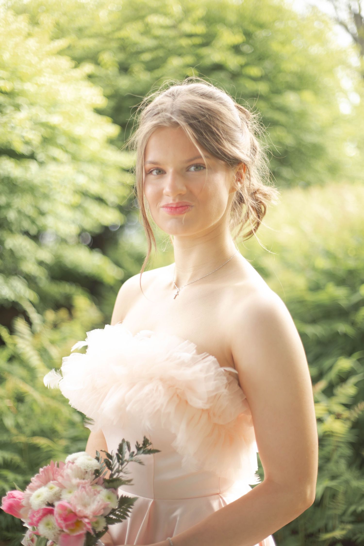 A figure in a blush pink strapless dress holds a bouquet against a soft green garden background.