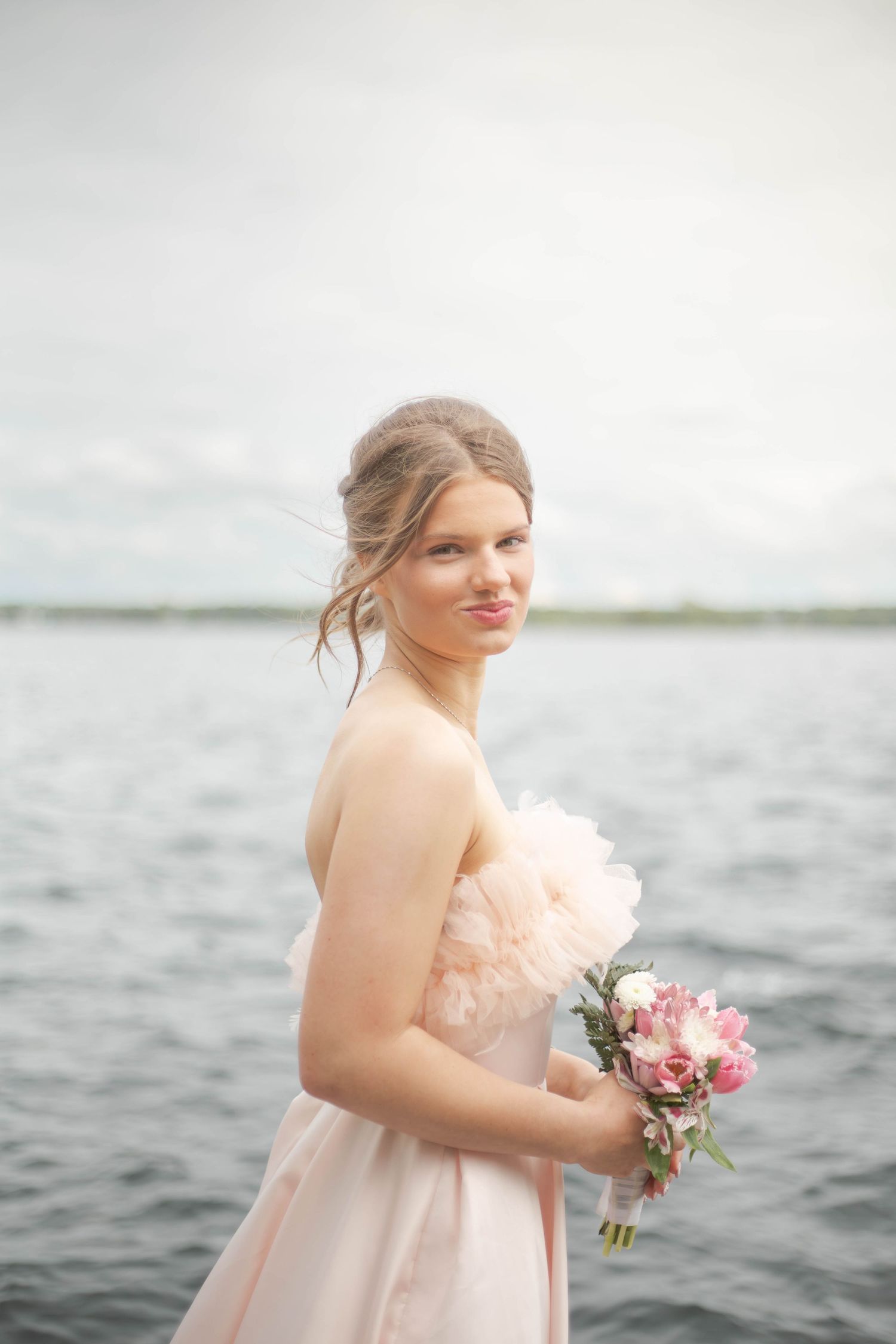Woman in blush pink dress holds pink floral bouquet while posing by waterfront on overcast day.