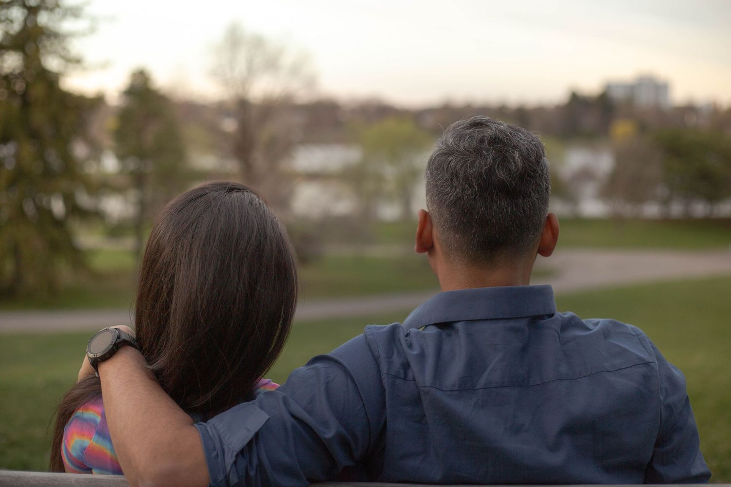 A couple sits close together outdoors at dusk, viewed from behind in a peaceful park setting.