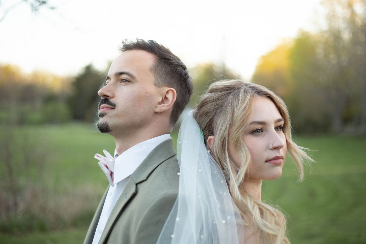 A joyful wedding couple poses together outdoors in a sun-lit green garden setting.