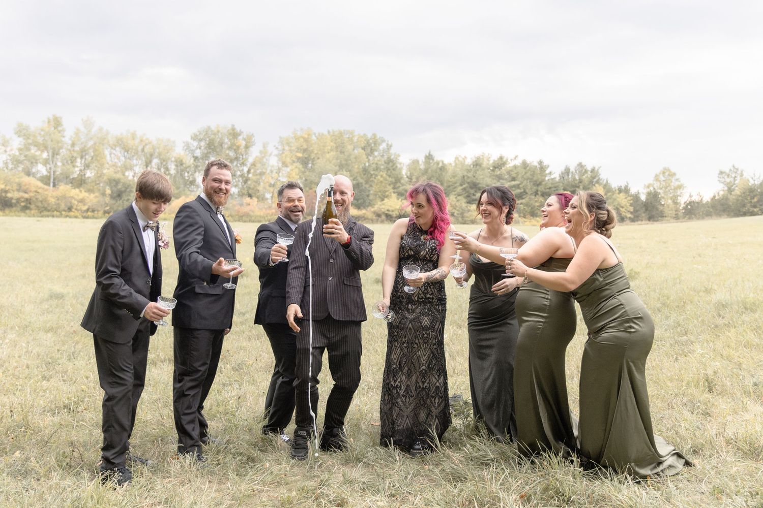 Wedding party laughs together while standing in a grassy field on a sunny autumn day.