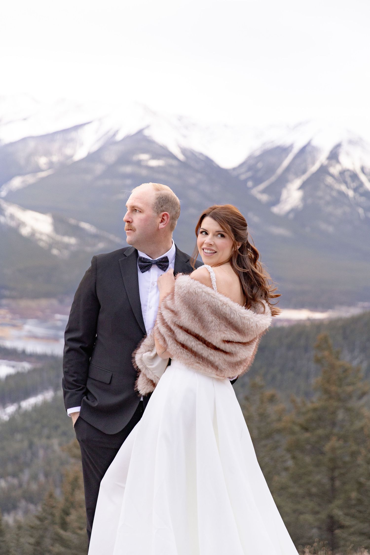 A bride in a white dress and beige shawl poses with her groom against a snowy mountain backdrop in winter.