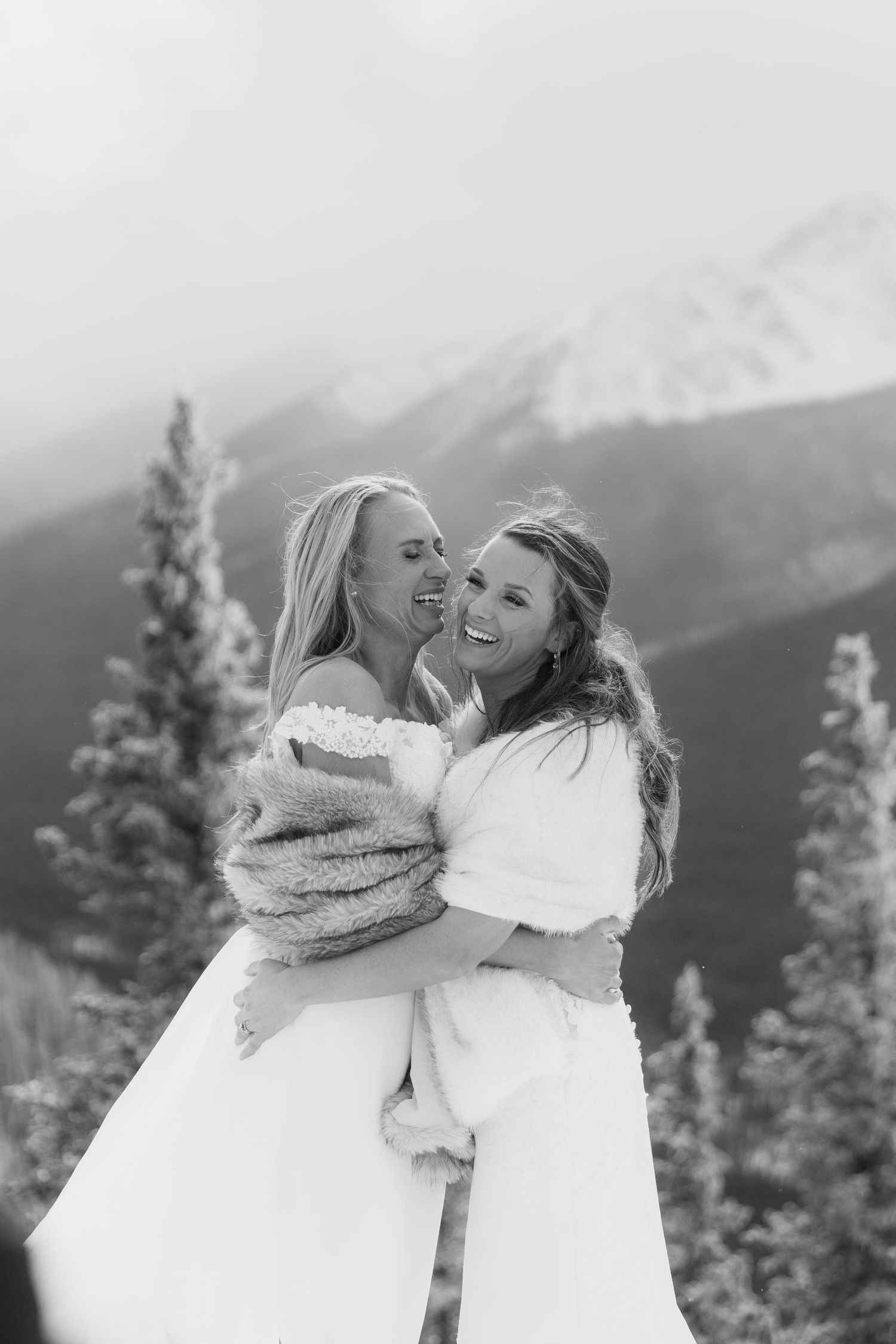 Two people share a joyful embrace against a backdrop of snow-capped mountains and evergreen trees in black and white.