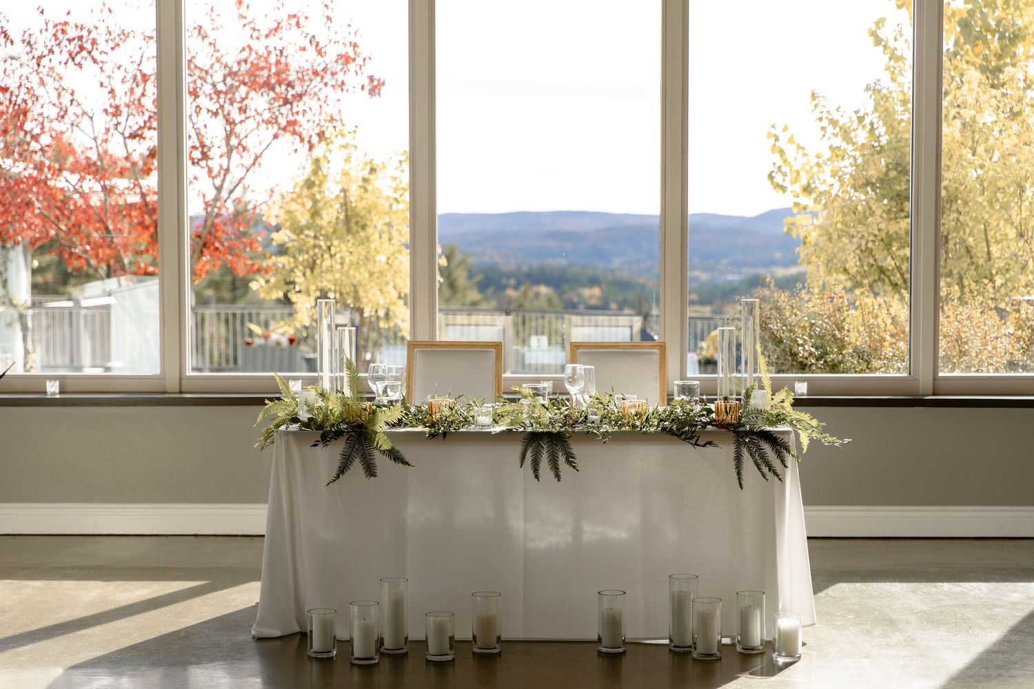 Elegant white-draped head table with floral garland decorations by large windows overlooking autumn trees and mountains.