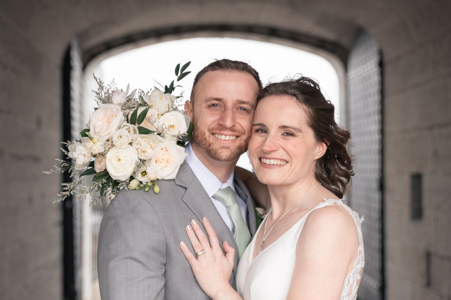 Happy bride and groom pose together holding white rose bouquet in front of stone arch on wedding day.