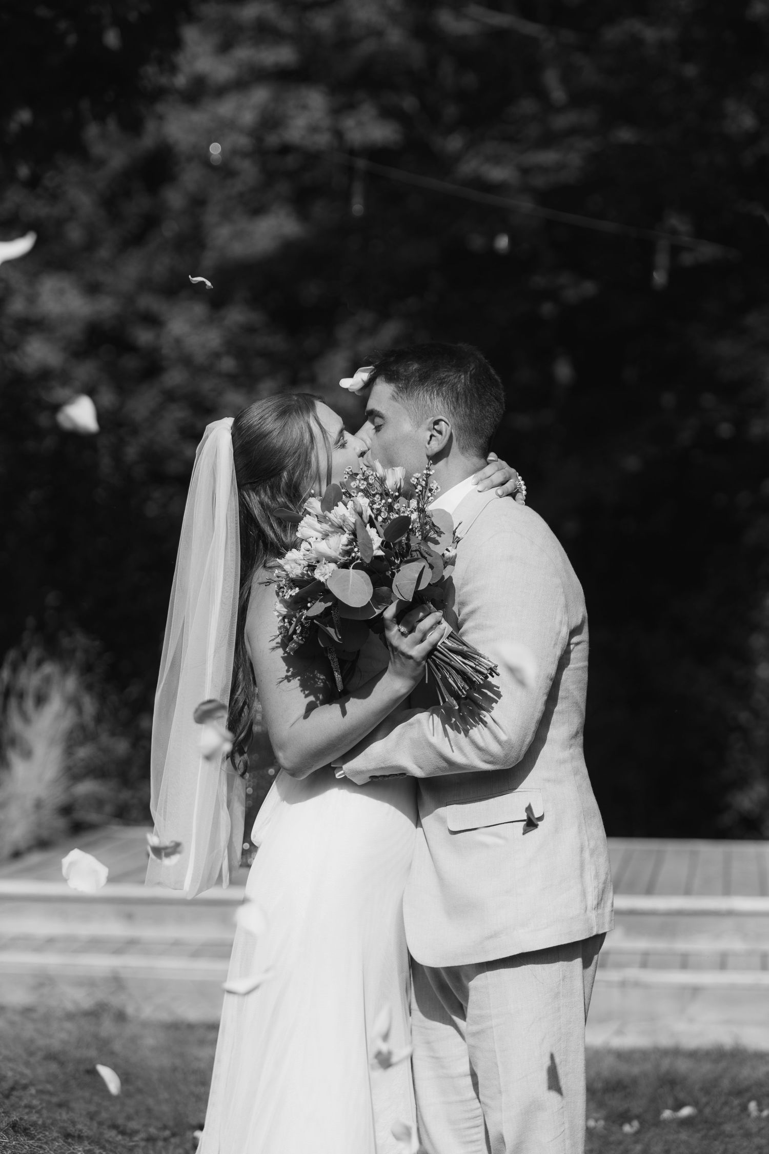 A romantic black and white wedding photo showing a couple sharing a kiss while holding a floral bouquet outdoors.