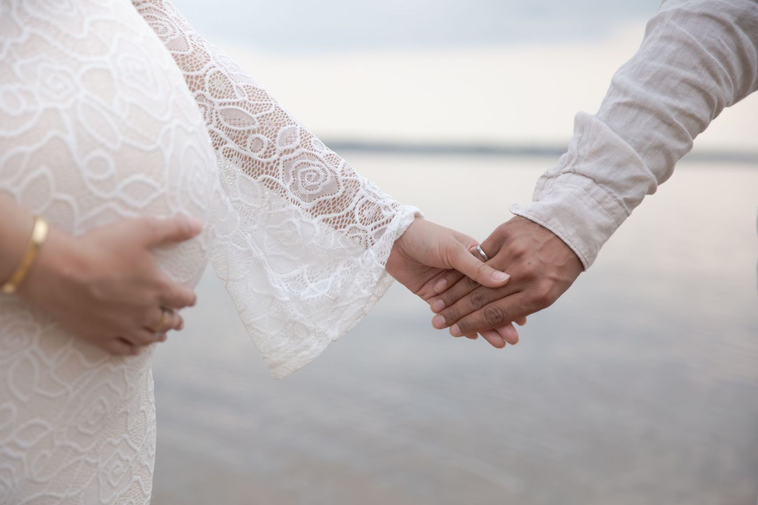 A couple holds hands tenderly on their wedding day, with a white lace dress visible by a tranquil waterfront.