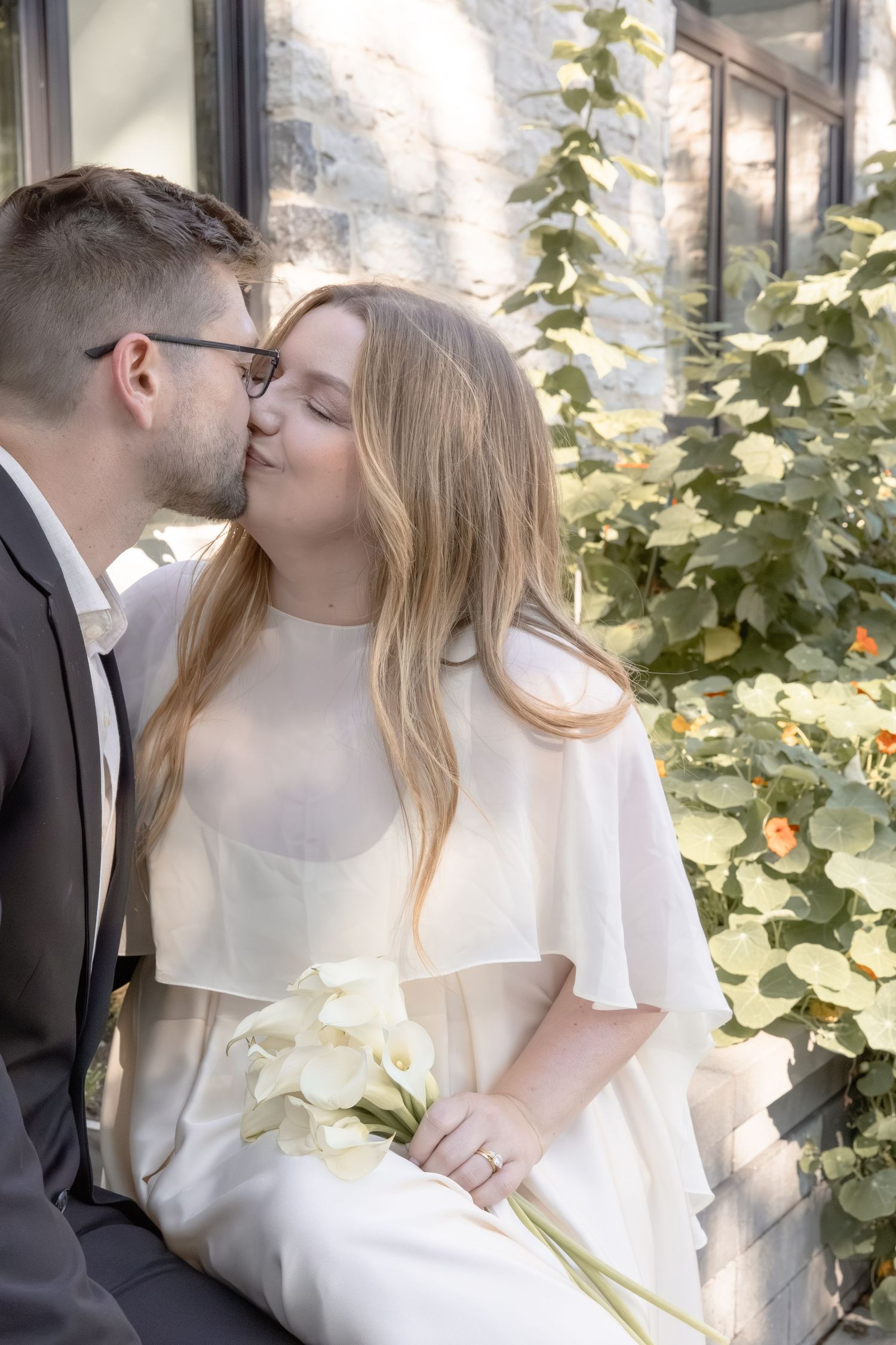 A bride and groom share romantic kisses in a garden setting with white roses and greenery.