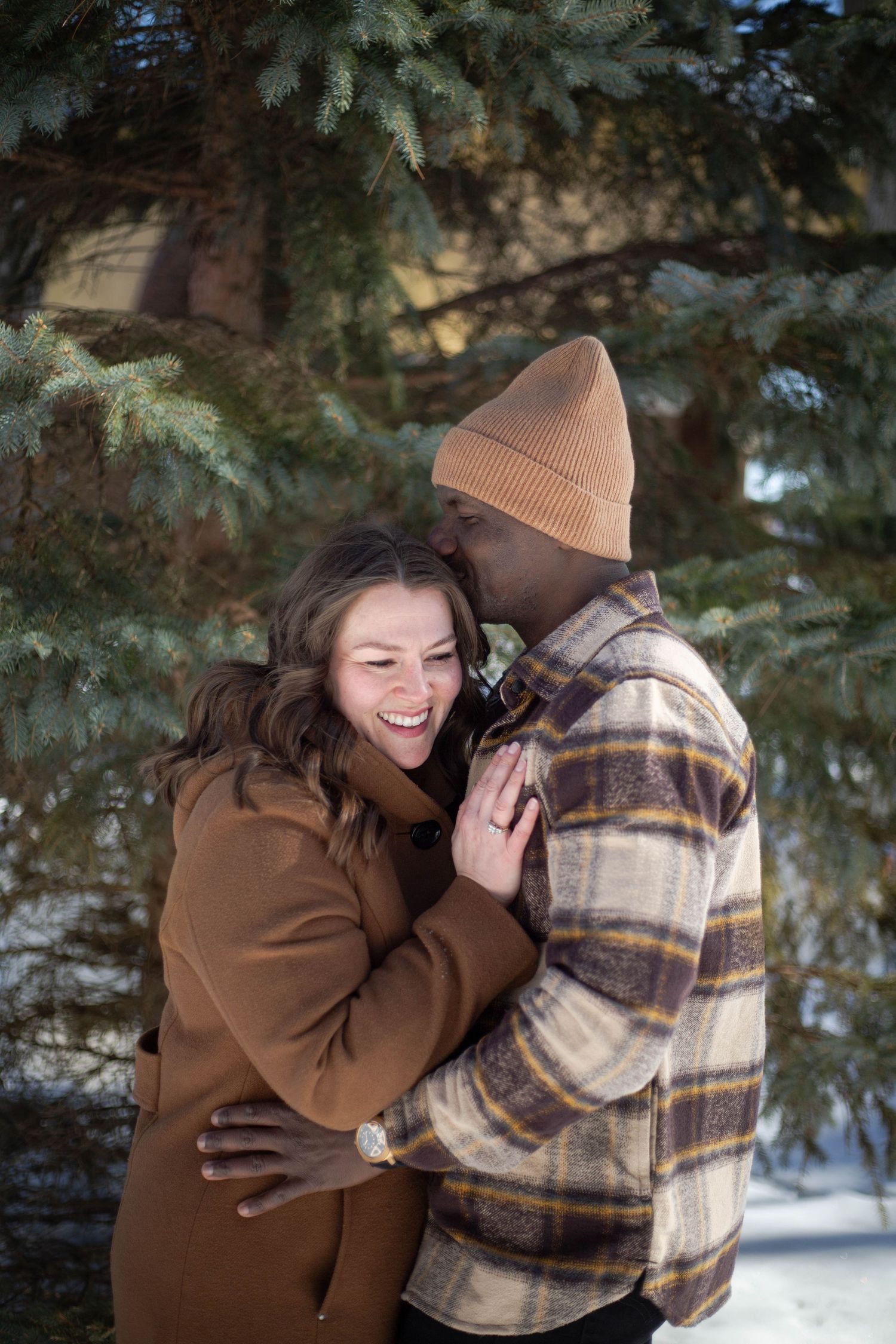 A couple shares a loving embrace and laugh together near evergreen trees in winter.