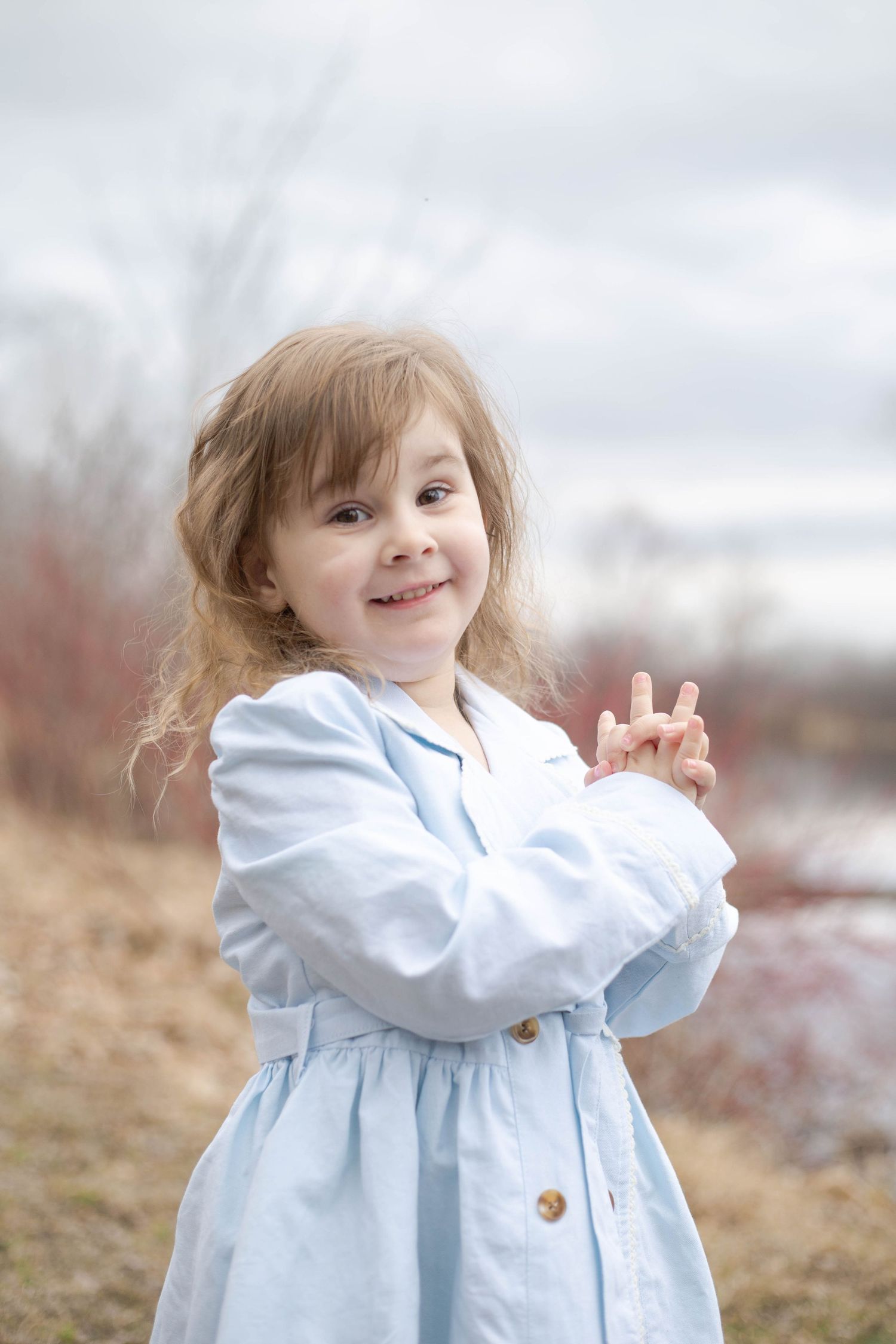 A young child in a light blue dress smiles outdoors on a cloudy day against a blurred natural background.