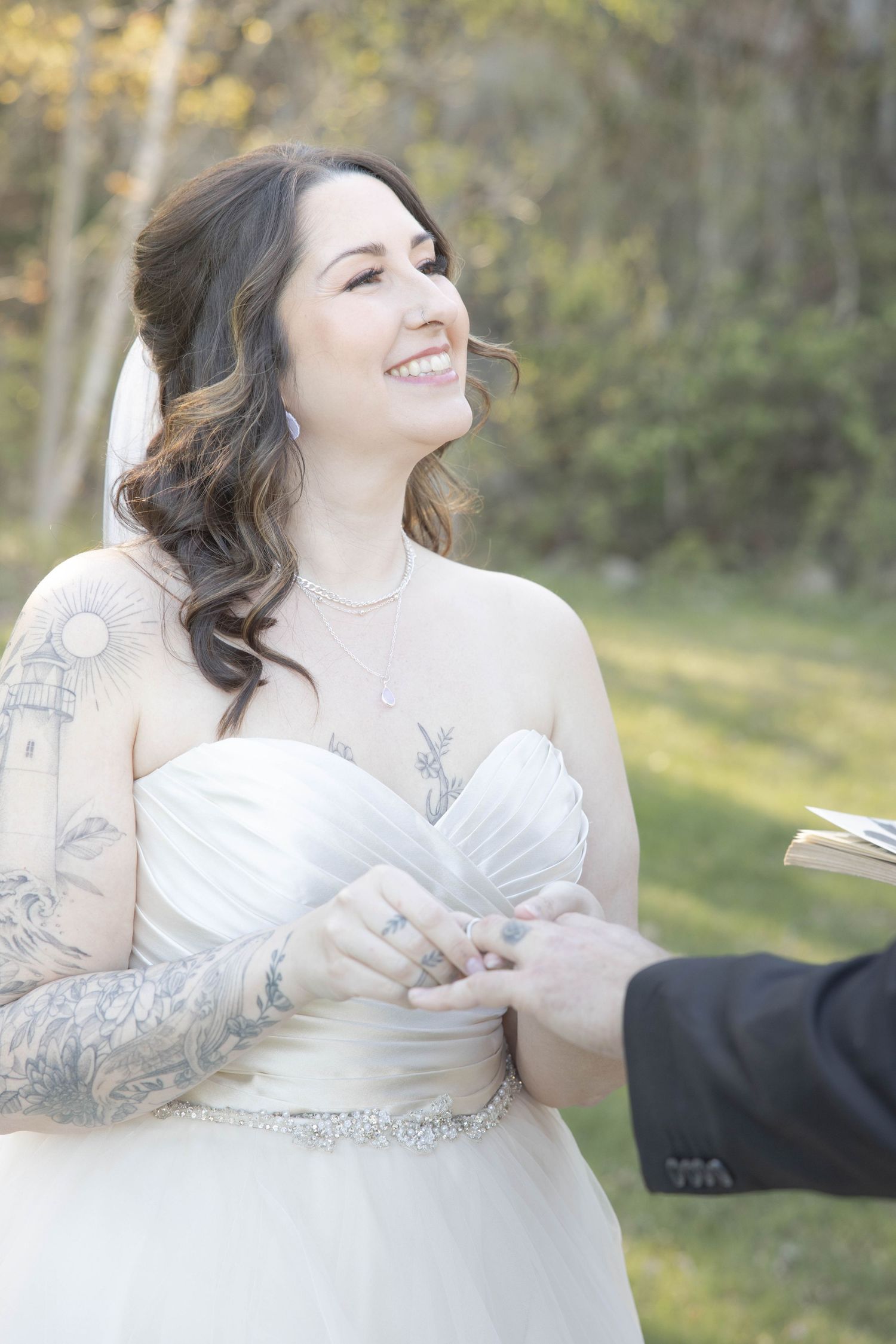 A bride in a white dress with tattoos smiles joyfully during an outdoor wedding ceremony in golden sunlight.