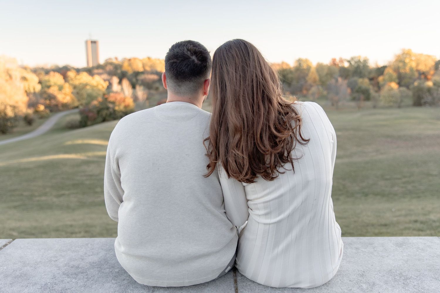 A couple in matching white sweaters sits together on a concrete ledge overlooking a grassy park at sunset.