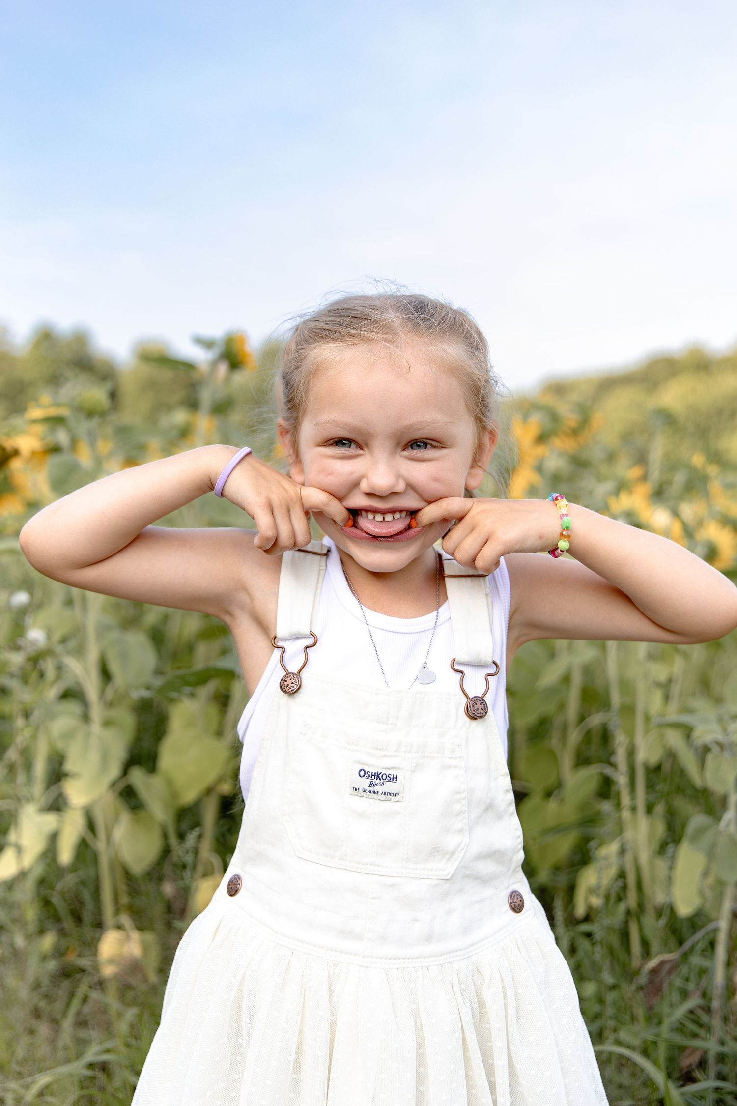 A cheerful child in white overalls makes a silly face while standing in a sunflower field on a sunny day.