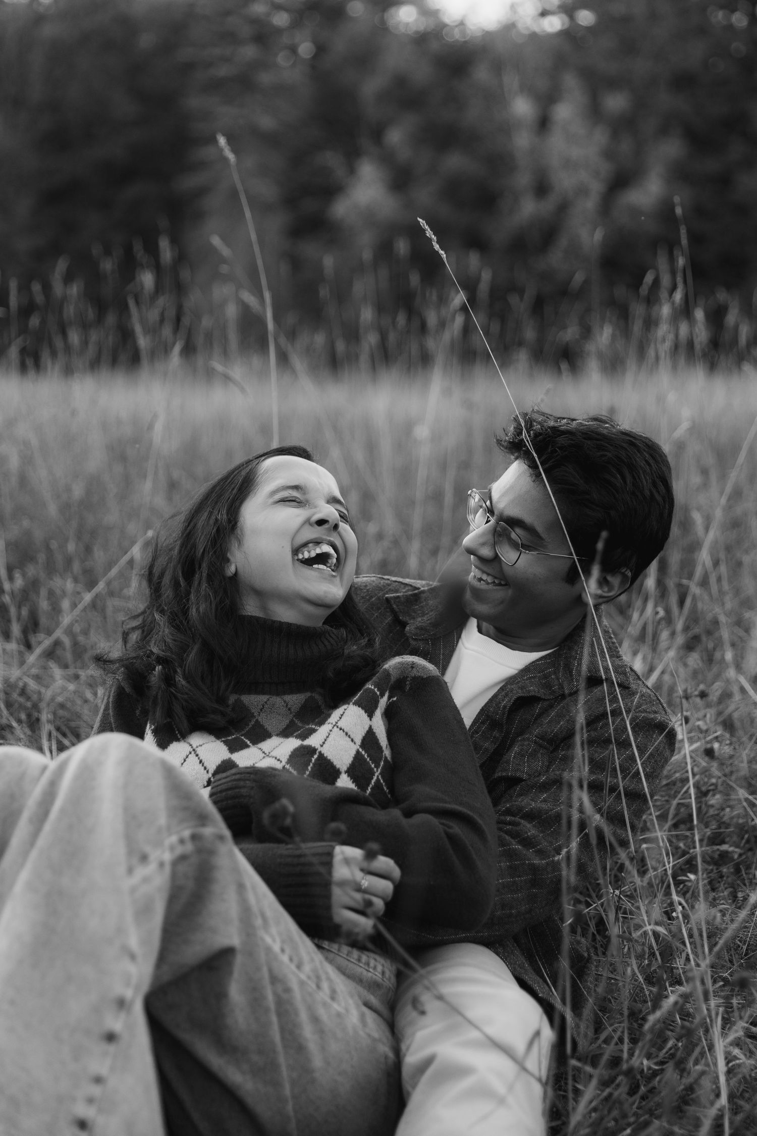 A black and white photo series shows two people laughing joyfully while lying in tall grass outdoors.
